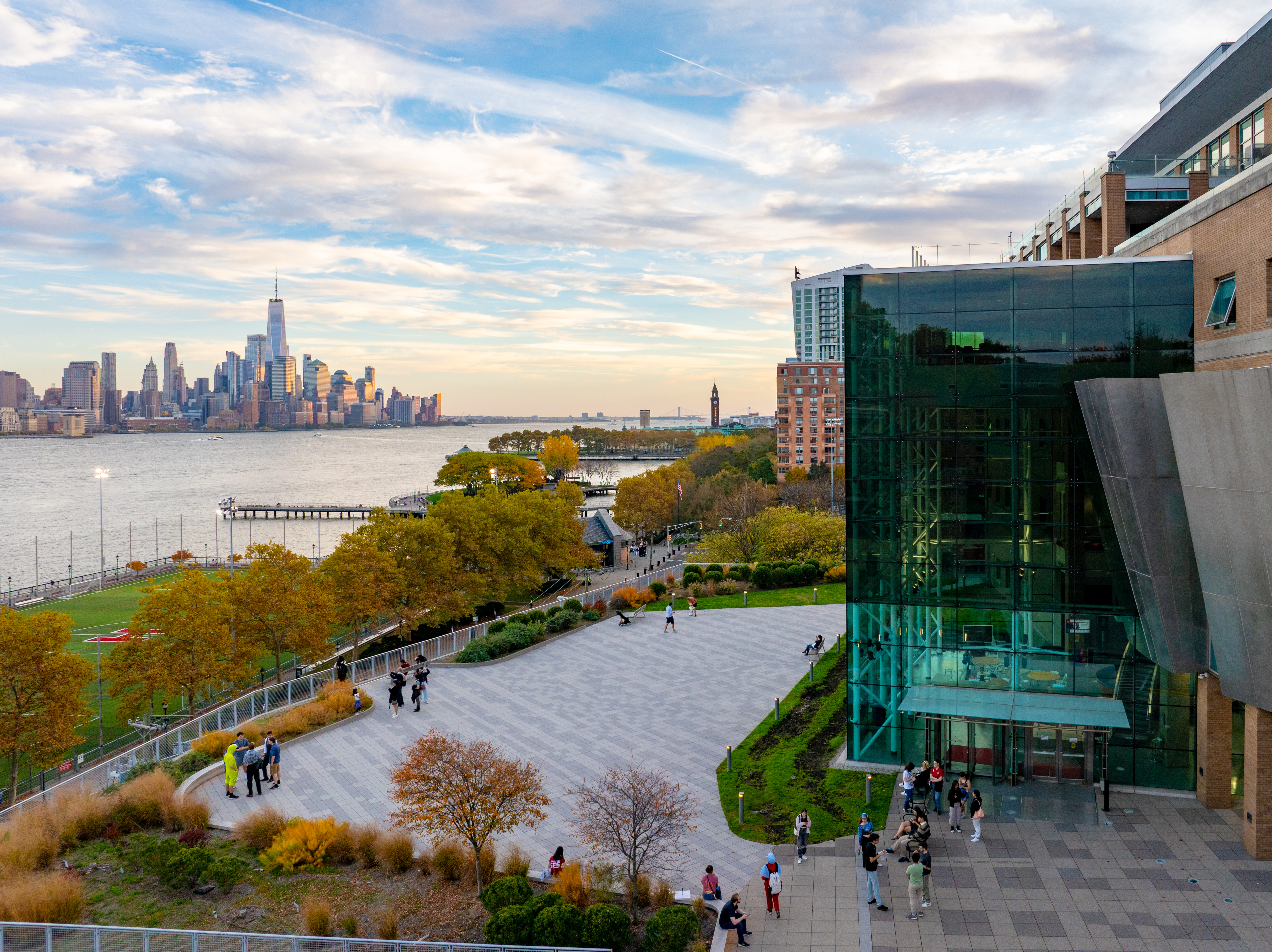 Babbio Center in the fall. Multiple groups of people stand around talking on the patio. There are fall trees. Part of the New York City skyline can be seen in the background.