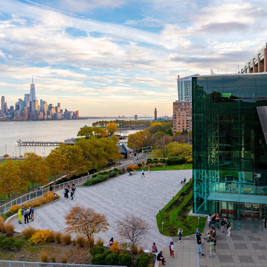 Babbio Center in the fall. Multiple groups of people stand around talking on the patio. There are fall trees. Part of the New York City skyline can be seen in the background.