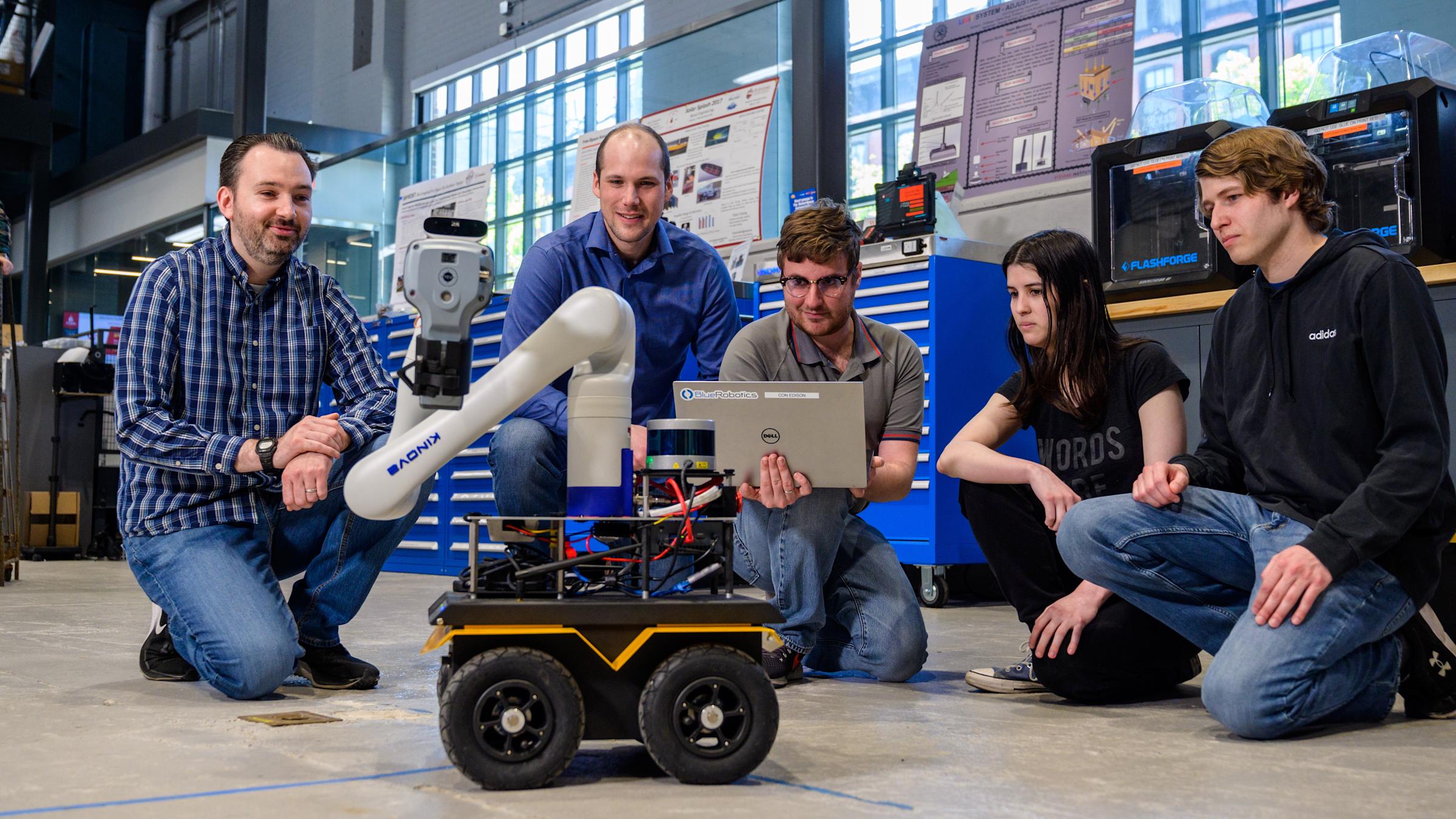 Professor Brendan Englot and a group of students using a laptop to operate a four wheeled robot with a crane neck/arm.