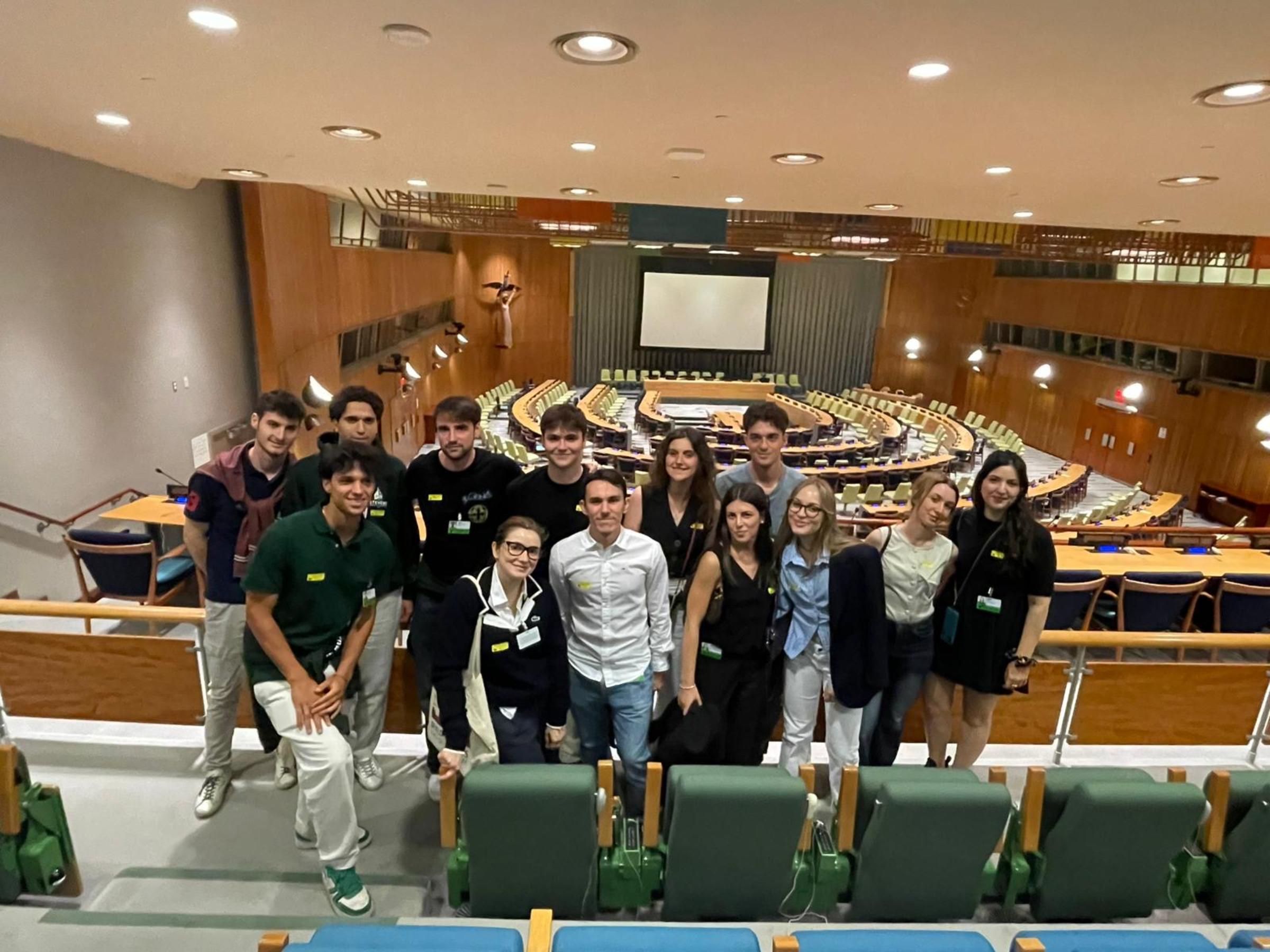 Students and their instructor pose inside the United Nations General Assembly chamber, standing in tiered seating with the iconic curved wooden desks, projection screen, and interpreter booths visible behind them.