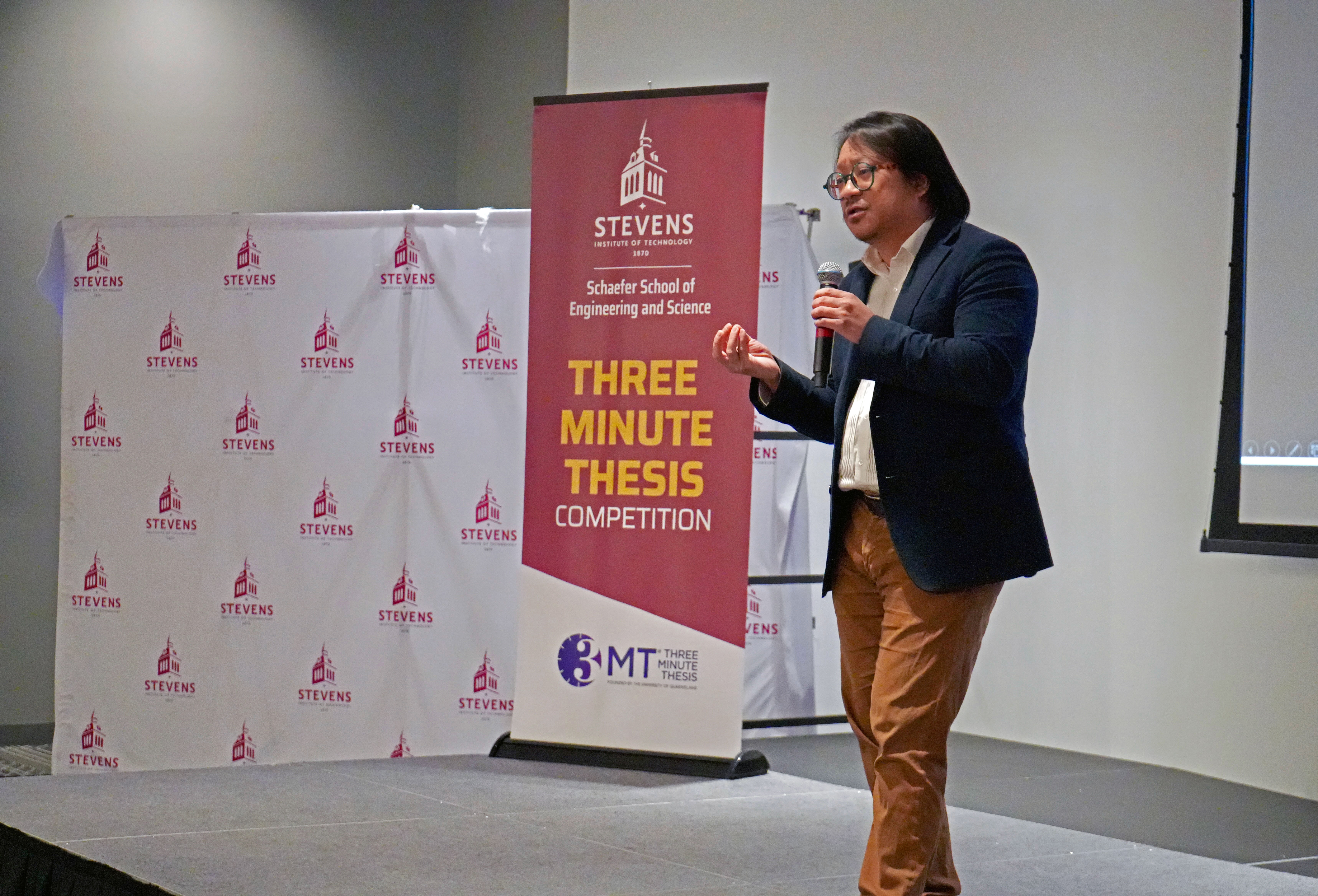 A student speaks on stage through a microphone with a Stevens Three-Minute Thesis Competition banner and backdrop.