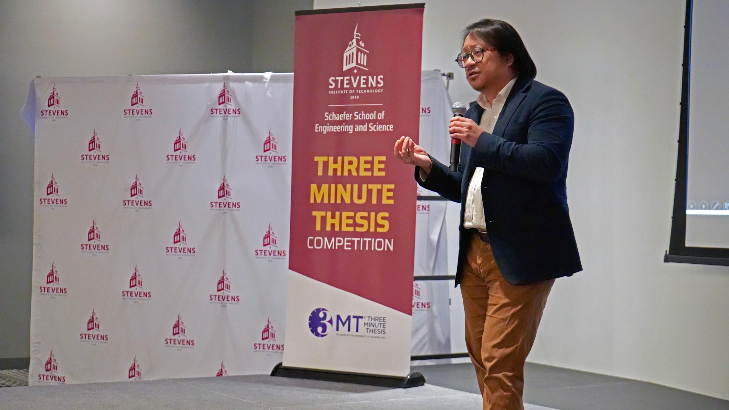 A student speaks on stage through a microphone with a Stevens Three-Minute Thesis Competition banner and backdrop.