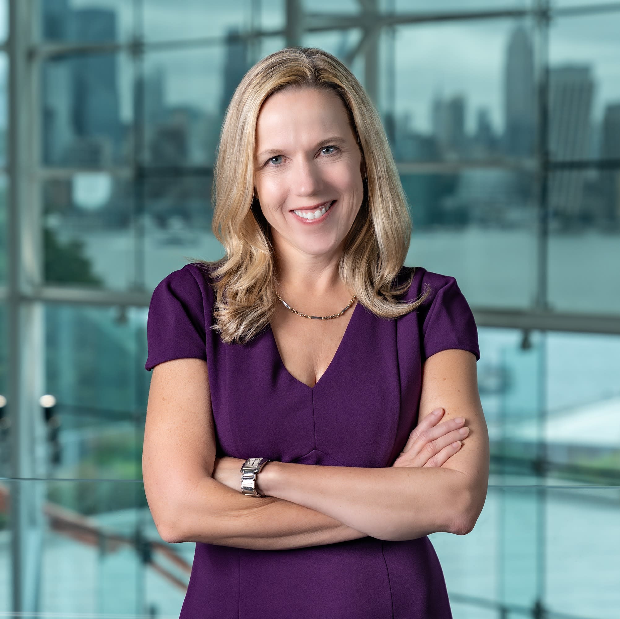 Professional woman with shoulder-length blonde hair wearing a purple dress stands with arms crossed, smiling at camera in a modern office building with glass walls and city views in background.