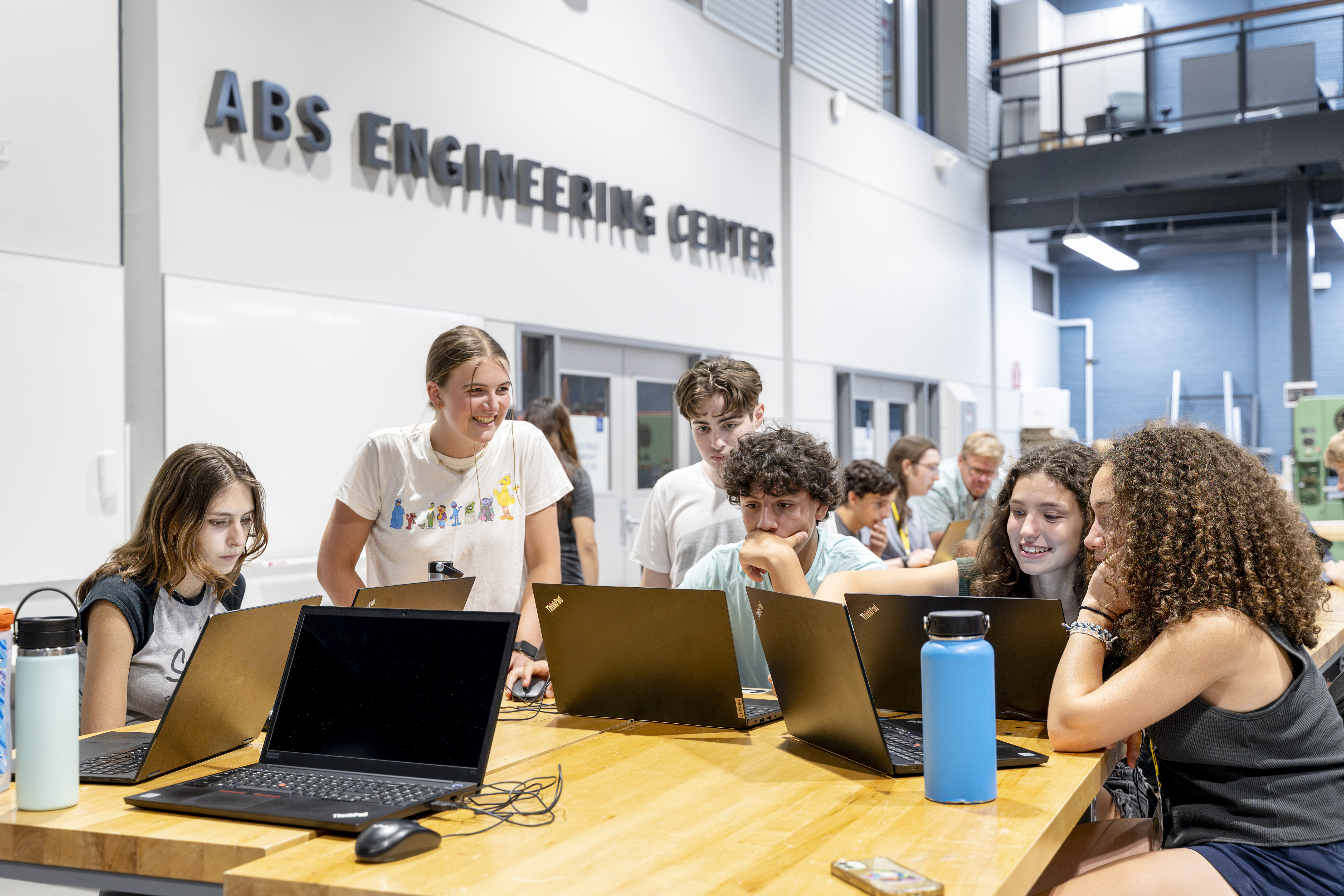 A group of six high school students, two boys and four girls, sit at a table in front of laptops, discussing a project in the ABS Engineering Center. 