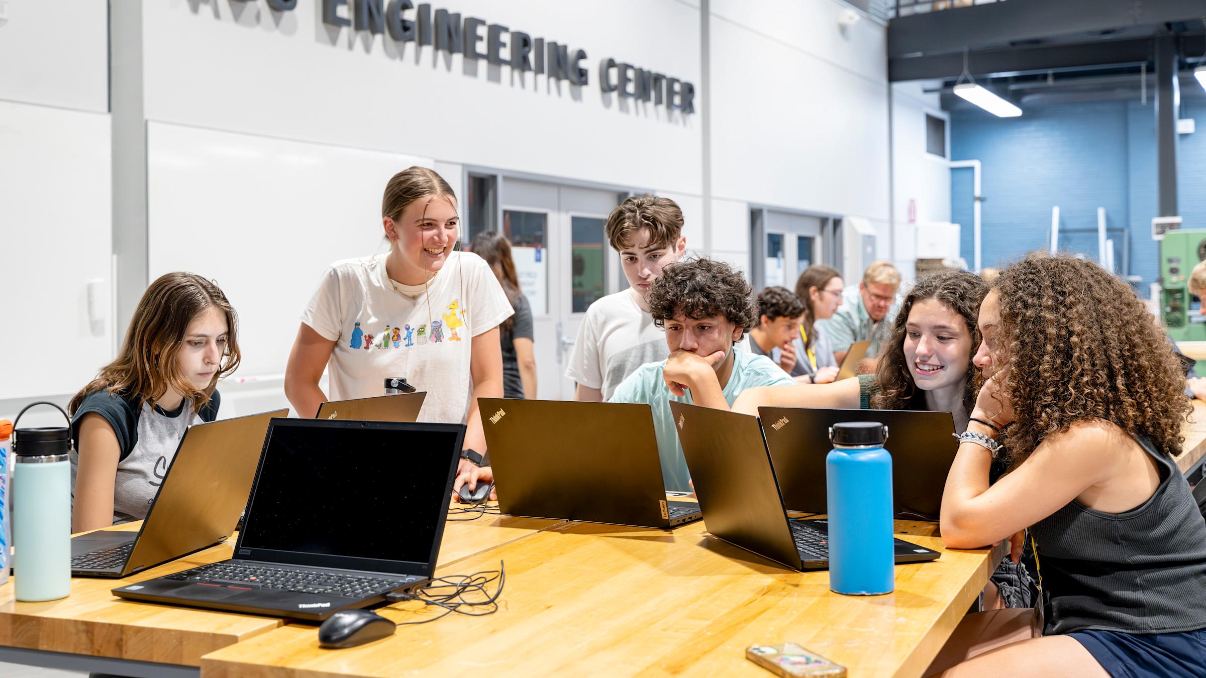 A group of six high school students, two boys and four girls, sit at a table in front of laptops, discussing a project in the ABS Engineering Center.