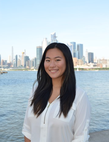 A photo of Katherine Ho standing before the Hudson River with the Manhattan skyline behind her