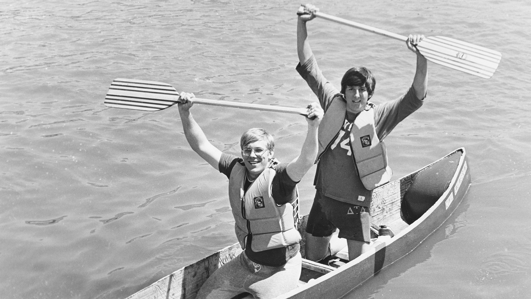 Two young men are in a canoe, one behind the other. The man in front is sitting, holding an oar in both hands over his head. The man in back is kneeling, also holding an oar in both hands over his head.