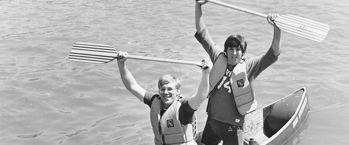 Two young men are in a canoe, one behind the other. The man in front is sitting, holding an oar in both hands over his head. The man in back is kneeling, also holding an oar in both hands over his head.