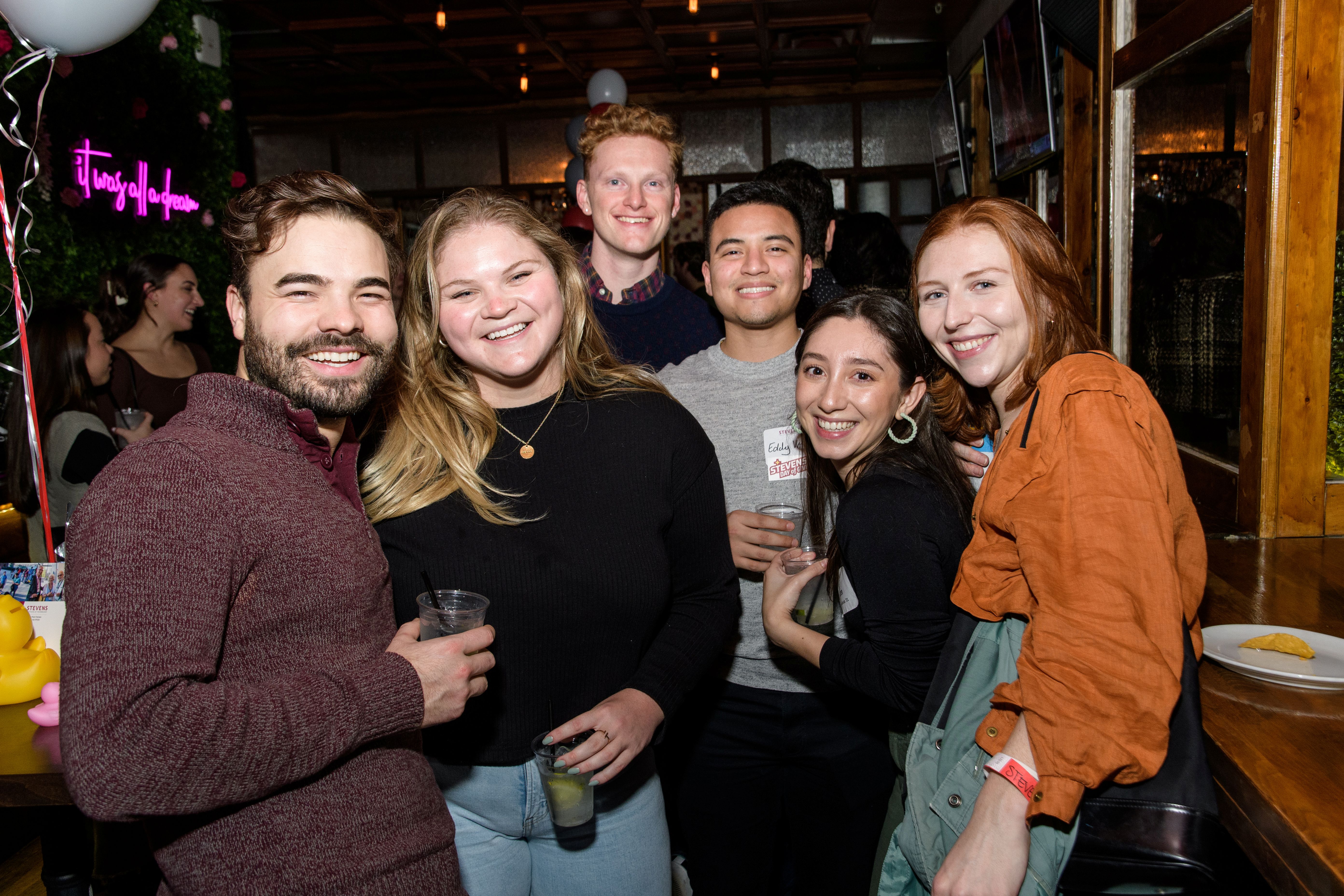 A group of six young alumni smile together holding drinks at a young alumni event 