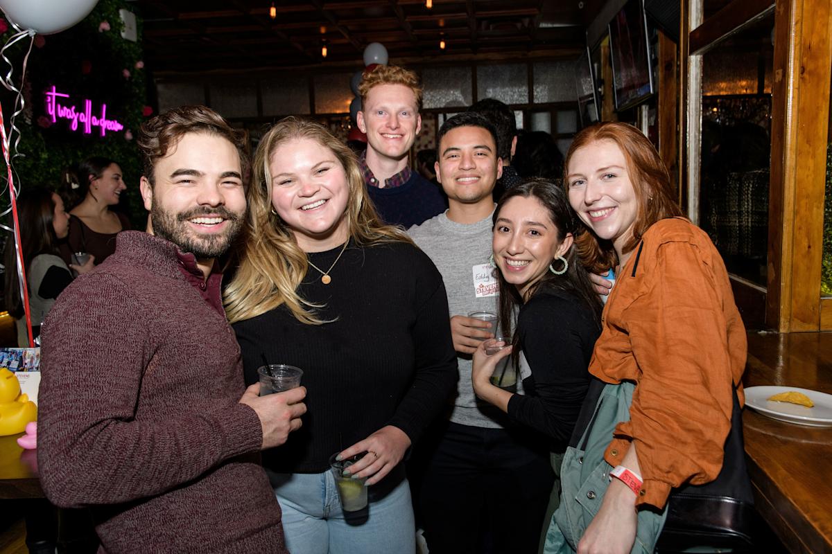 A group of six young alumni smile together holding drinks at a young alumni event 