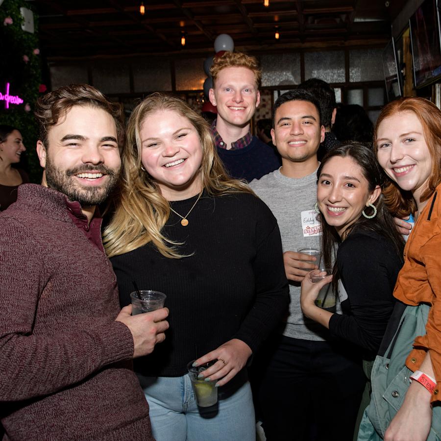 A group of six young alumni smile together holding drinks at a young alumni event