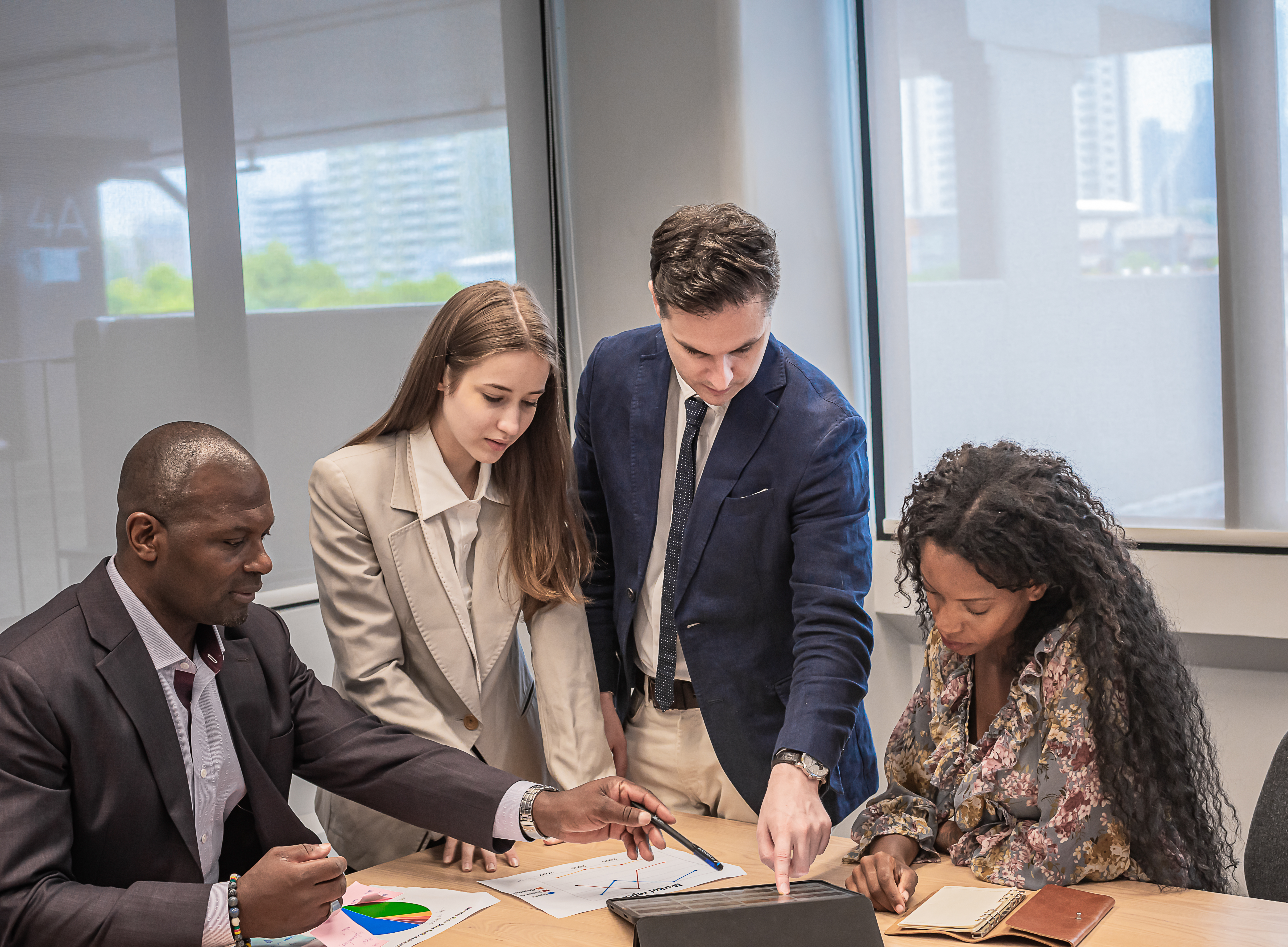 Four business professionals in suits collaborate around a conference table, reviewing documents and charts. They're gathered in a modern office with large windows showing a city view in the background.