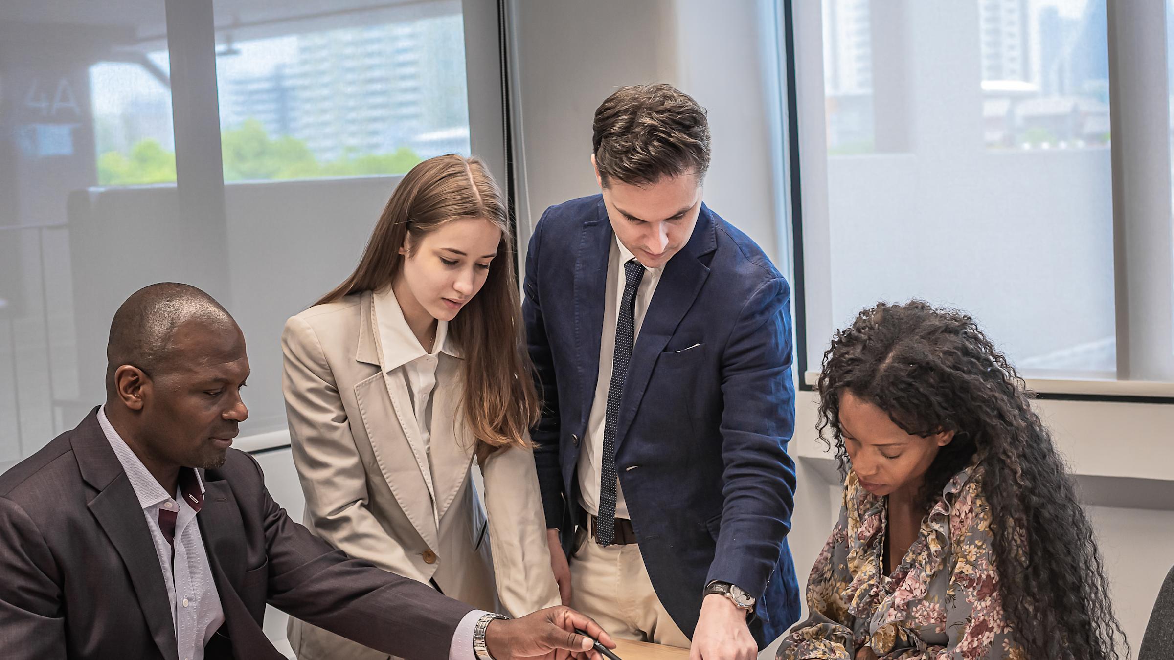 Four business professionals in suits collaborate around a conference table, reviewing documents and charts. They're gathered in a modern office with large windows showing a city view in the background.