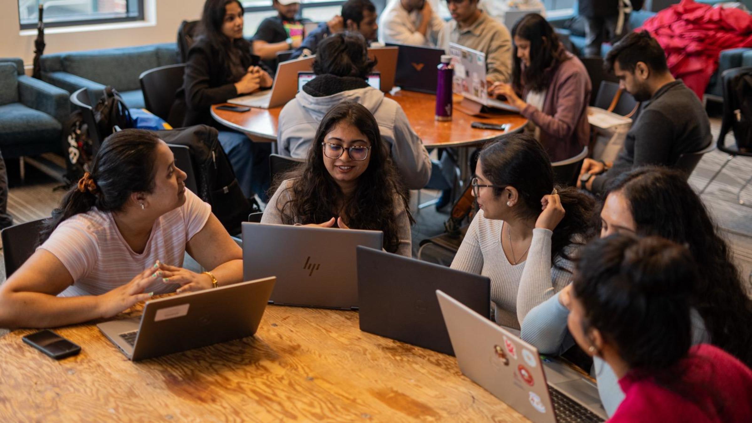 A group of female students sit around a table with their laptops collaborating with one another.