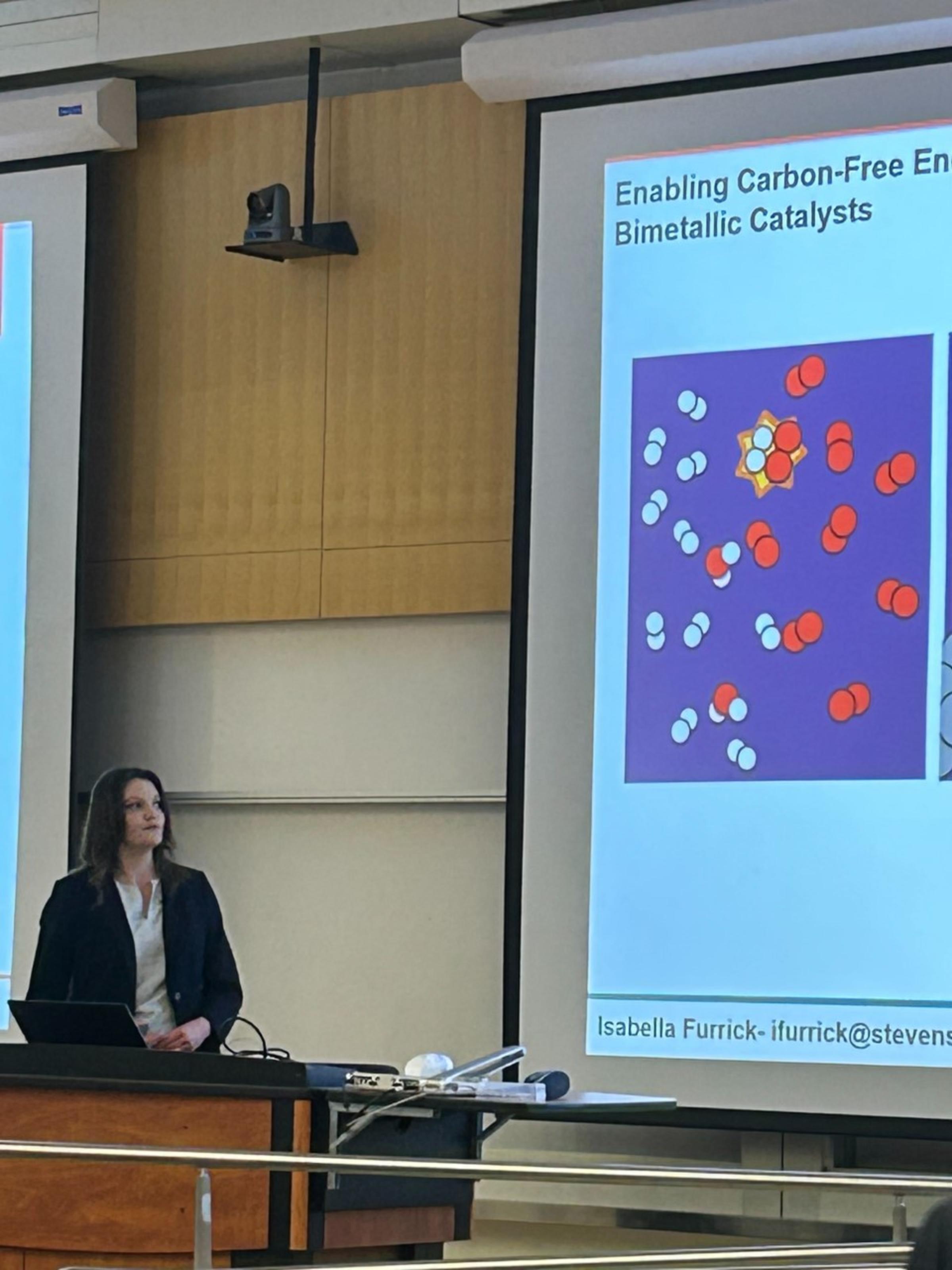 Photo of Isabella Furrick presenting her research, standing behind a podium and looking at research data on a large projector