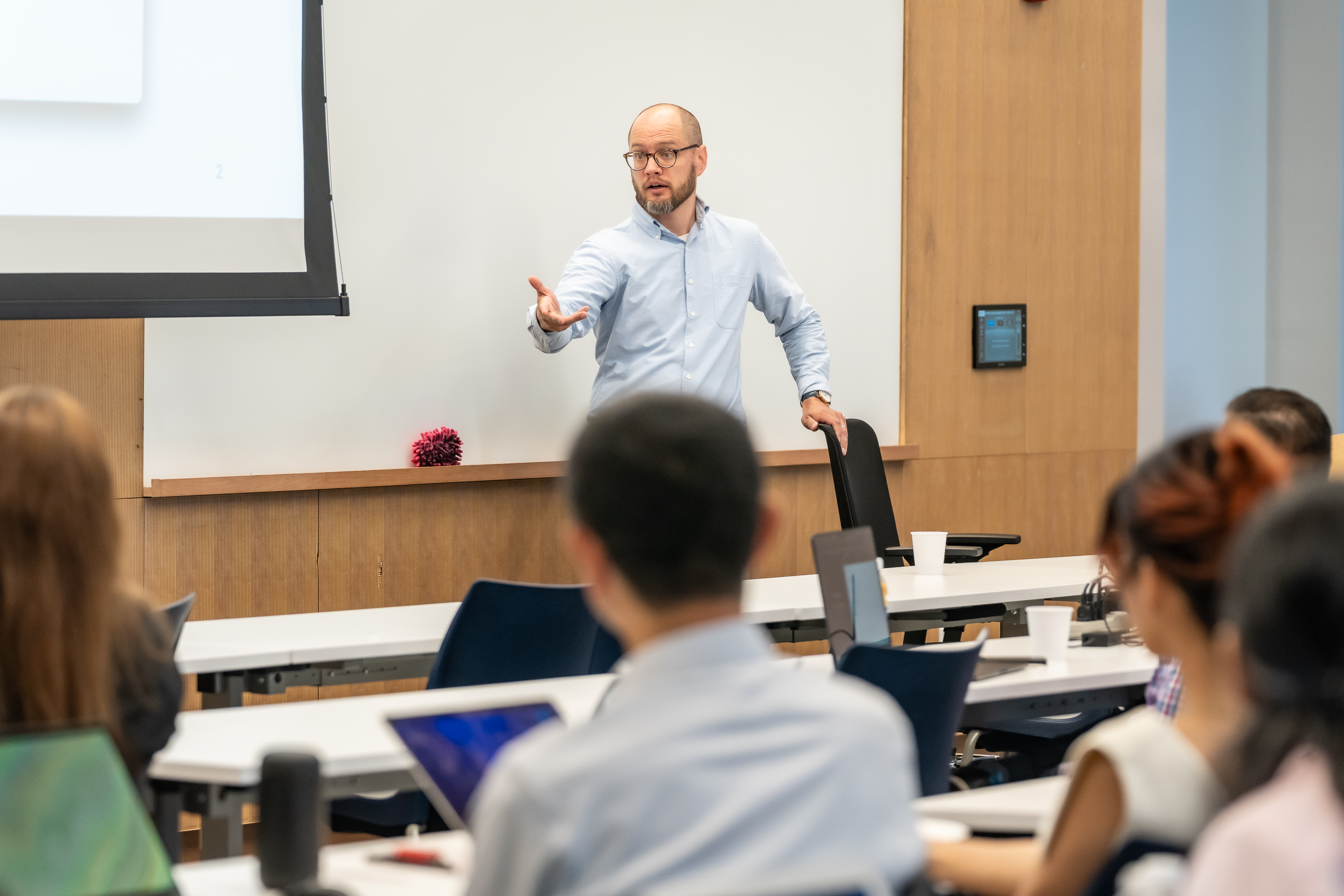 A bald man with glasses and beard in a light blue shirt presents to students in a modern lecture hall. He gestures while speaking from the front, with a projection screen behind him and students seated at tiered desks with laptops.