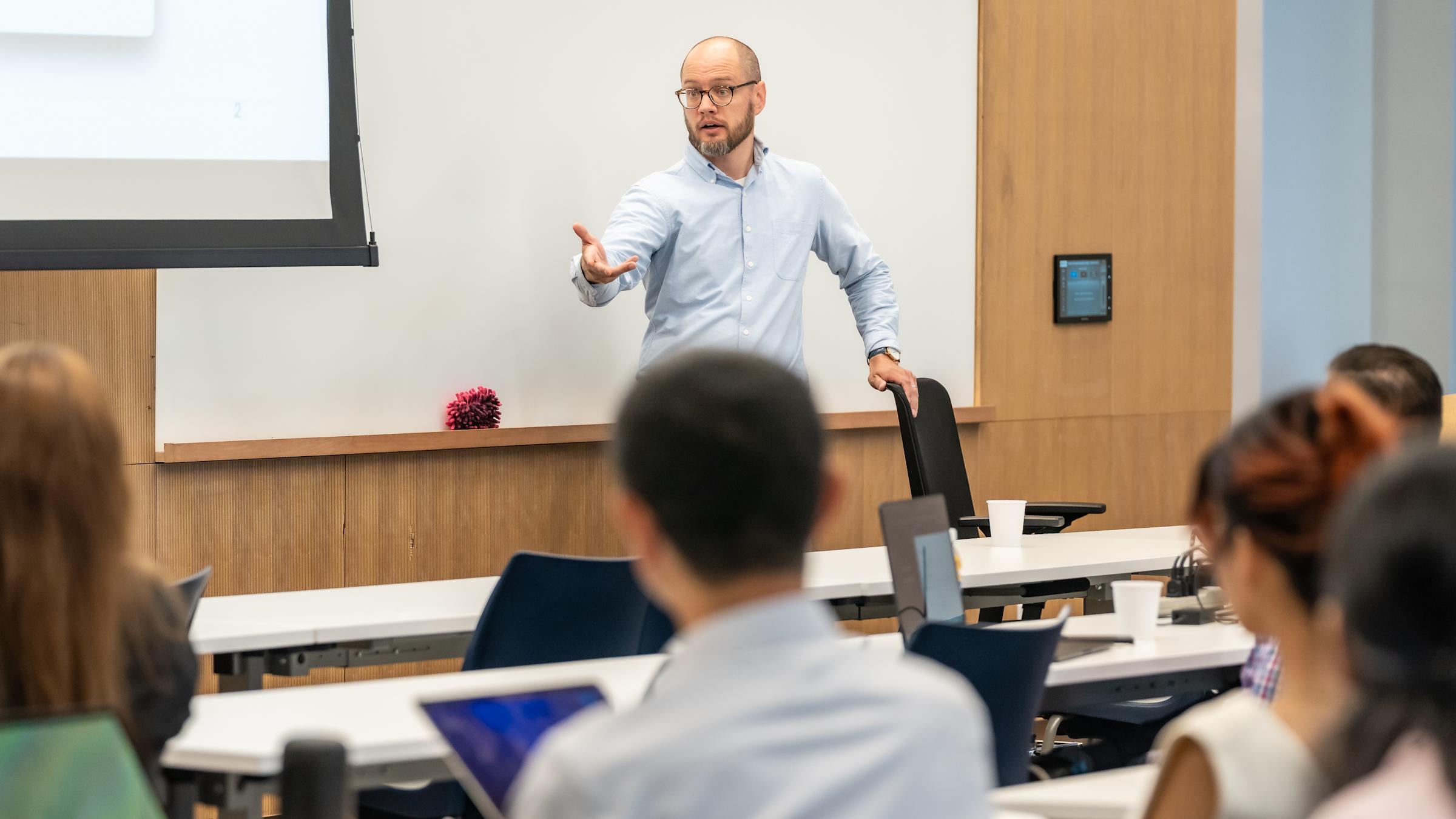 A bald man with glasses and beard in a light blue shirt presents to students in a modern lecture hall. He gestures while speaking from the front, with a projection screen behind him and students seated at tiered desks with laptops.