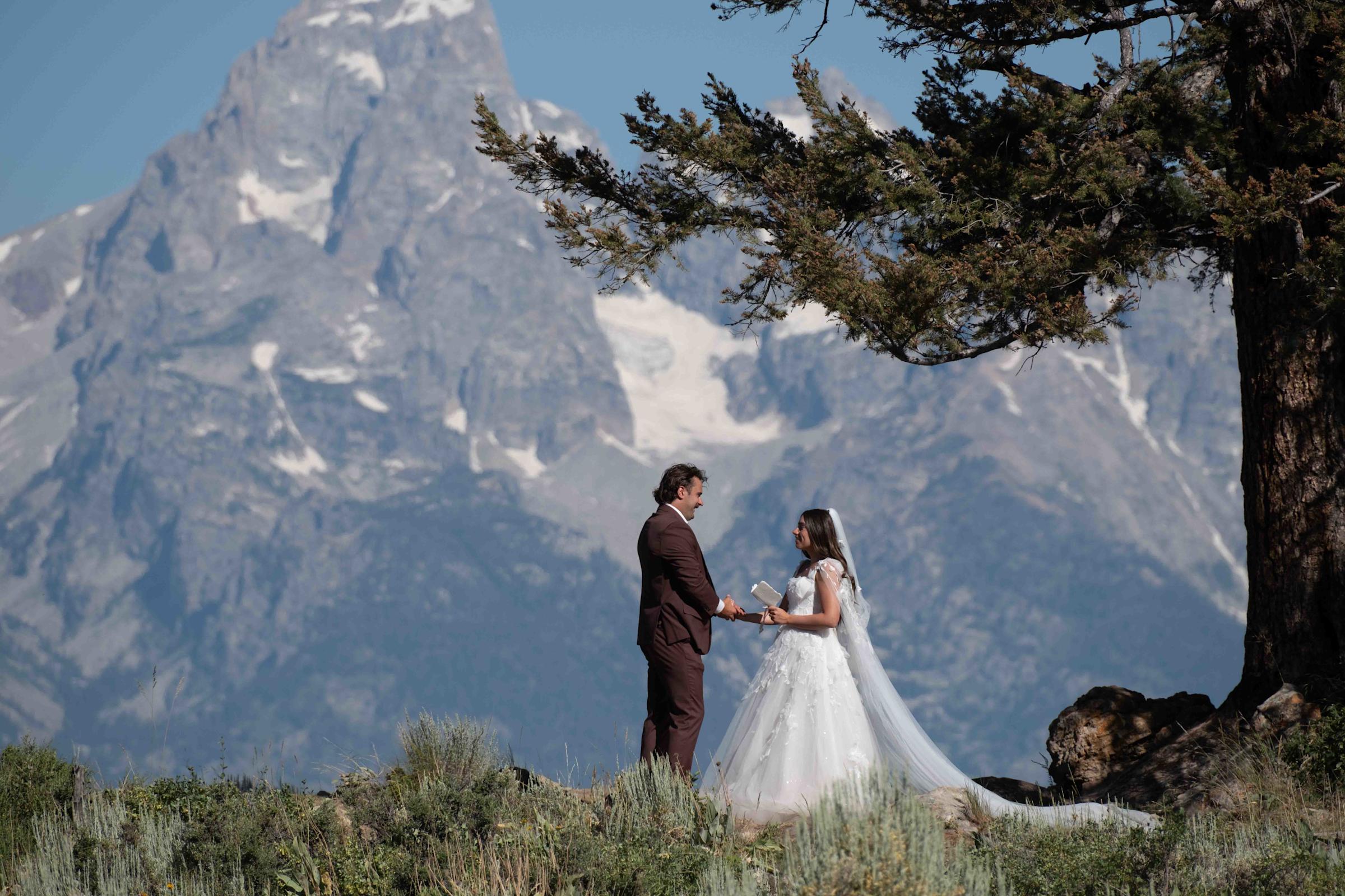 A couple exchanges vows during an outdoor wedding ceremony beneath a pine tree, with the dramatic snow-capped Grand Teton peak rising behind them. The bride wears a flowing white gown and veil; the groom wears a dark burgundy suit.