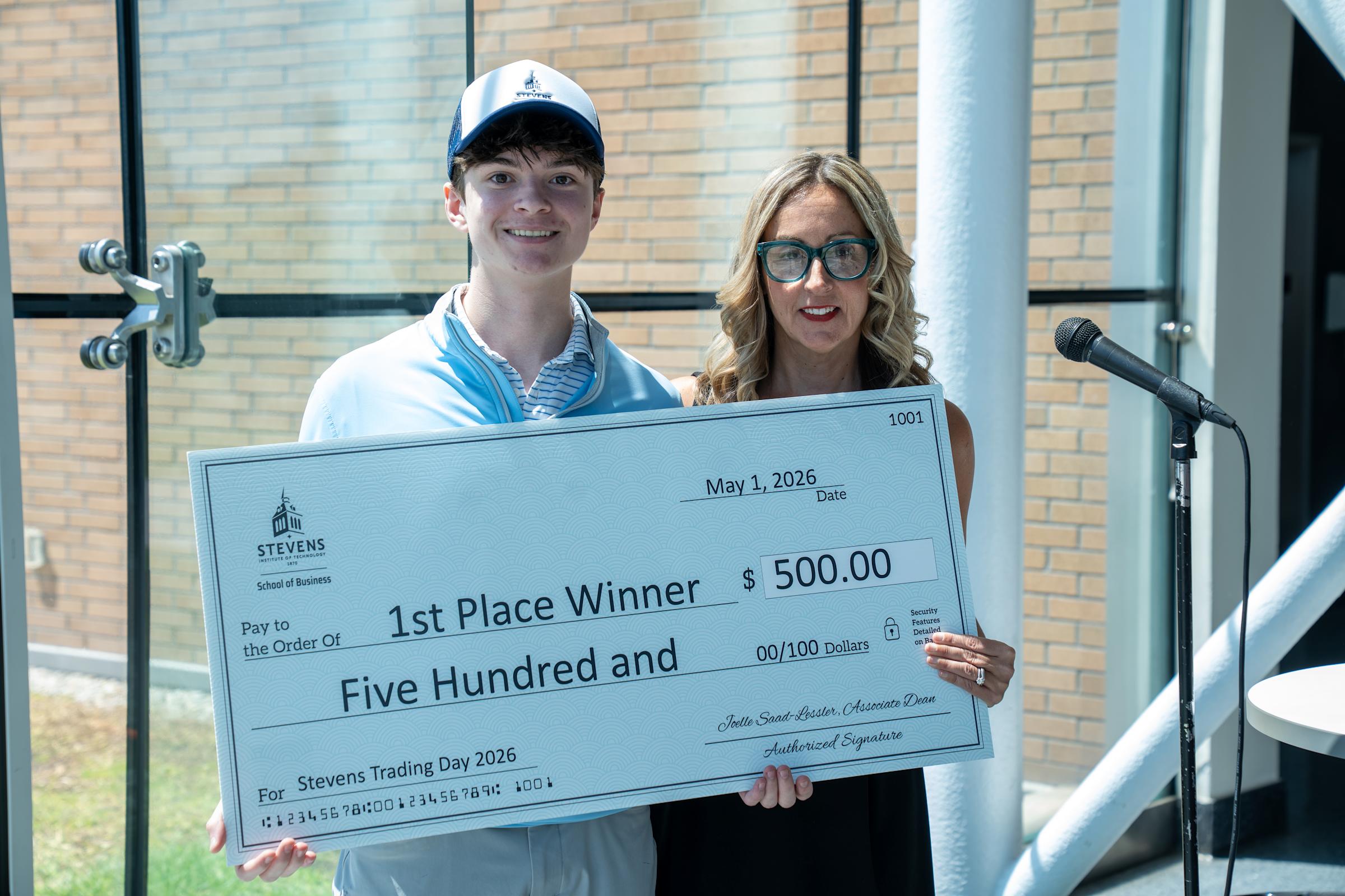 A young male student wearing a white Stevens baseball cap and light blue jacket poses with a woman in black and teal glasses holding a large ceremonial check for $500 made out to "1st Place Winner" from Stevens School of Business, dated May 1, 2026.