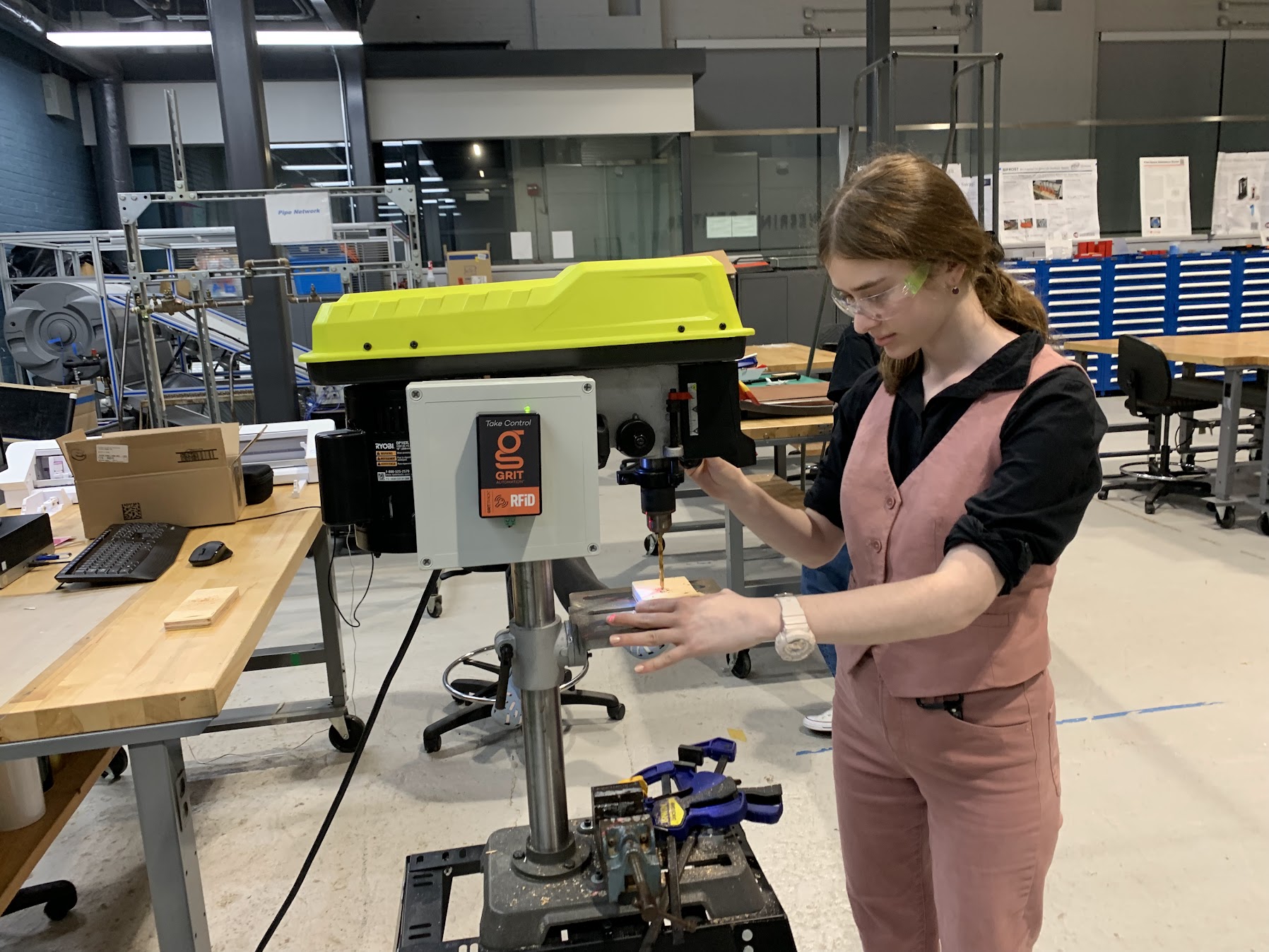 A female student wearing goggles stands in front of a machine she is operating, while holding an object in her hands that she is building.