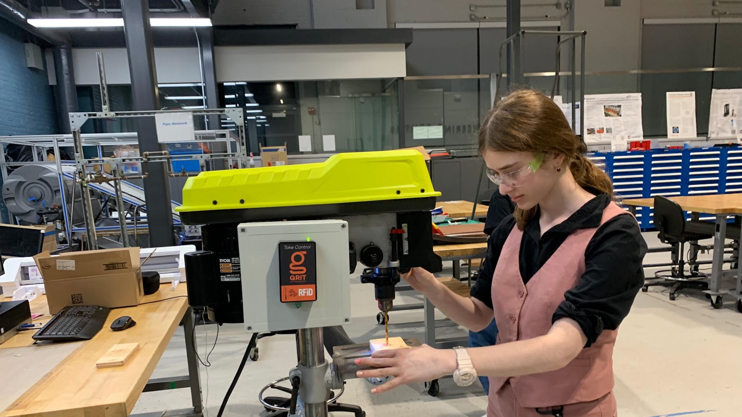 A female student wearing goggles stands in front of a machine she is operating, while holding an object in her hands that she is building.