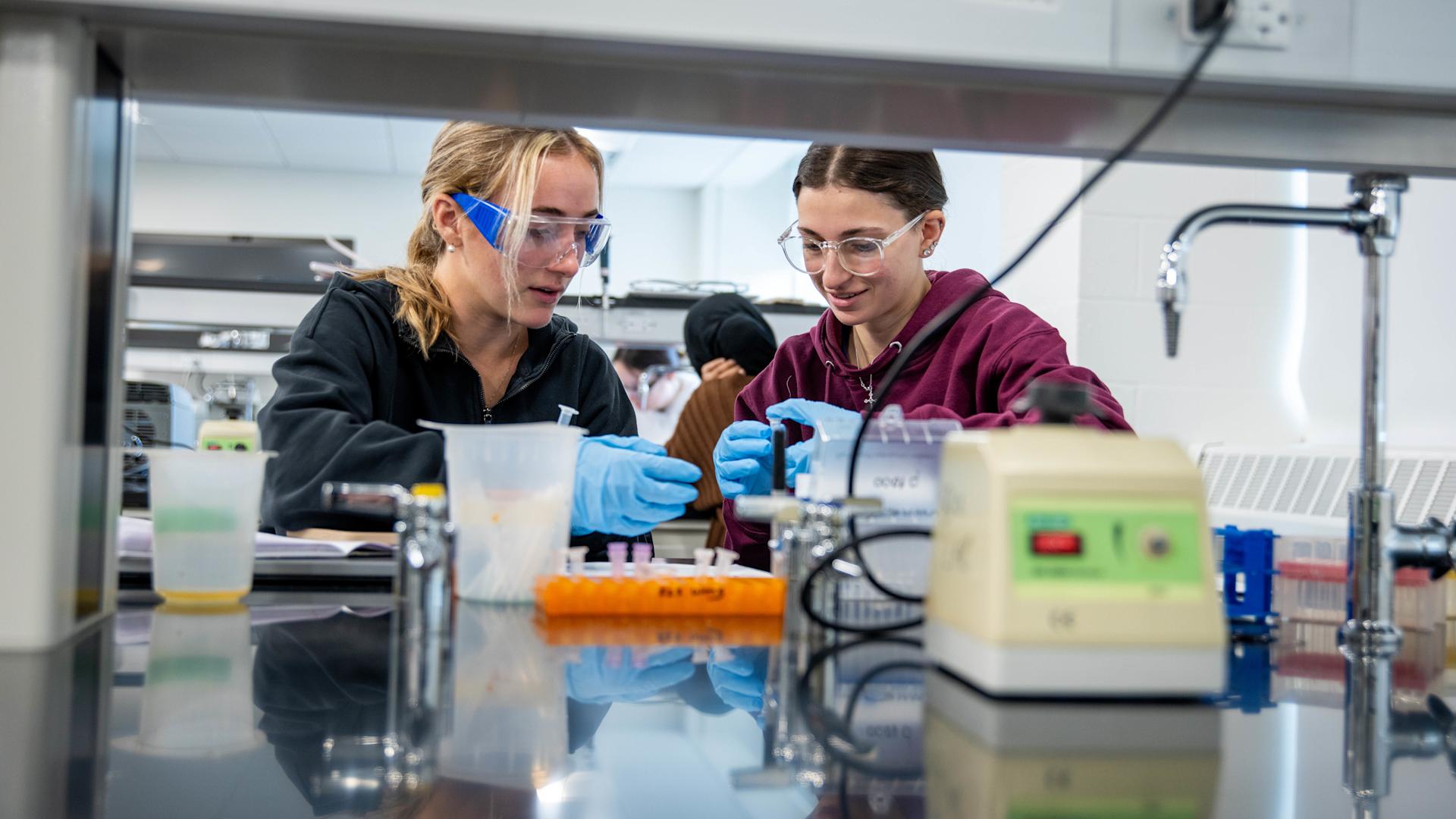 Two female students use medical equipment, including vials, in a lab setting.