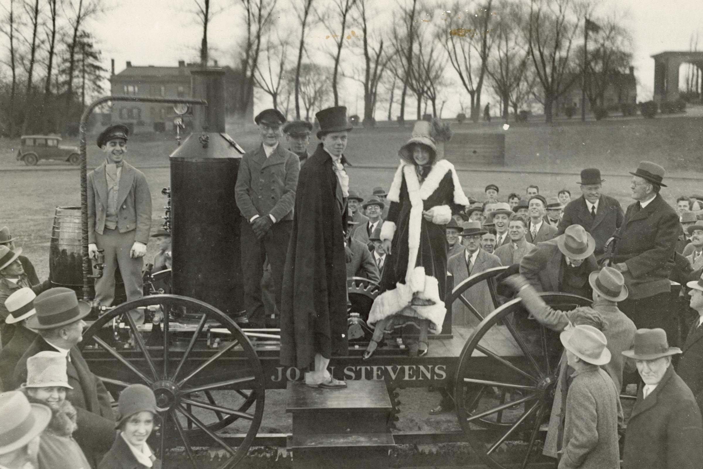 Black and white photo of a crowd around an old steam locamotive with a man in a top hat and a woman in a winter coat aboard.