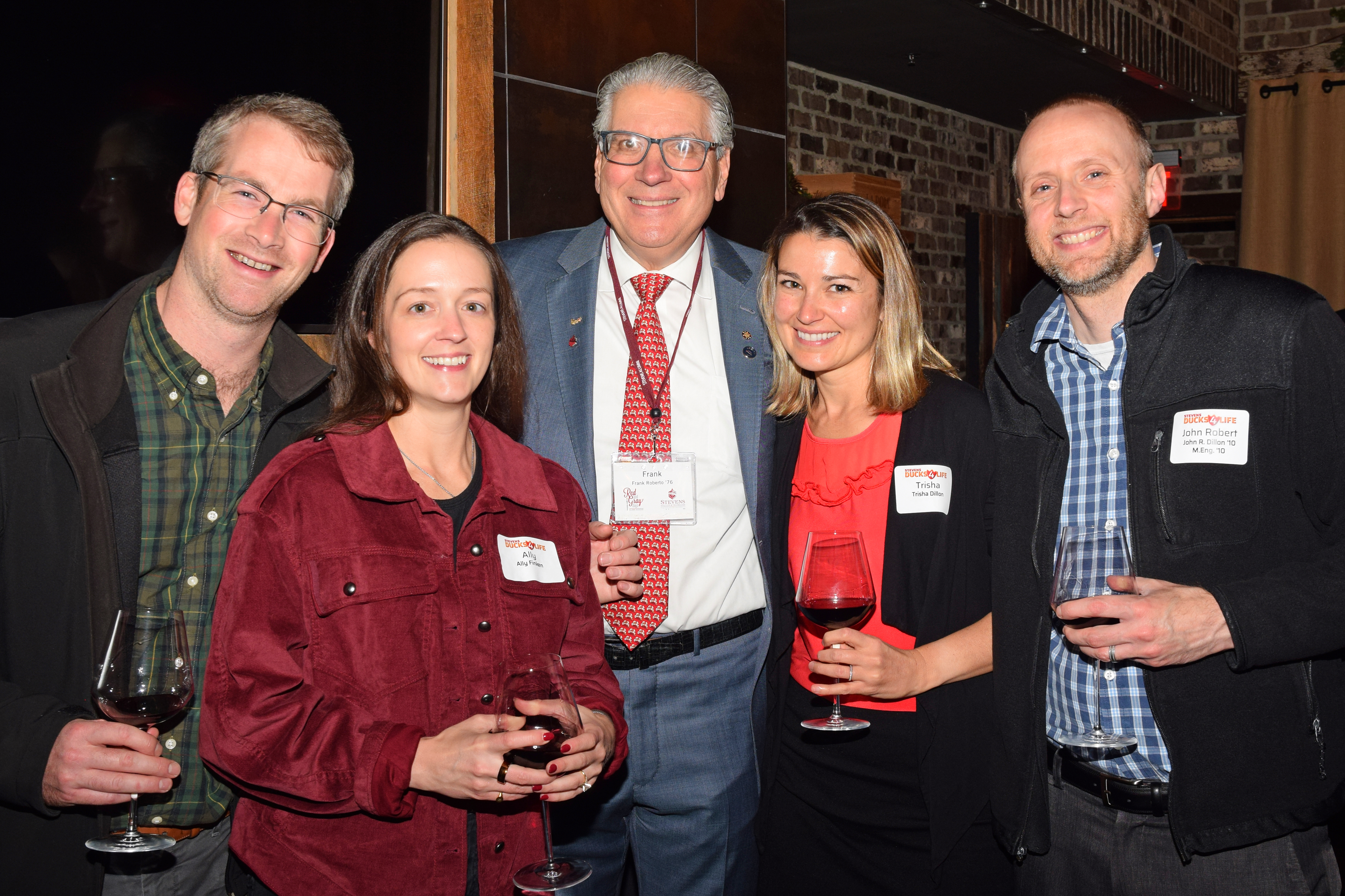 Five attendees pose for a photo at the Houston Alumni Holiday Gathering 2025, holding wine glasses in a warmly lit venue with brick walls.