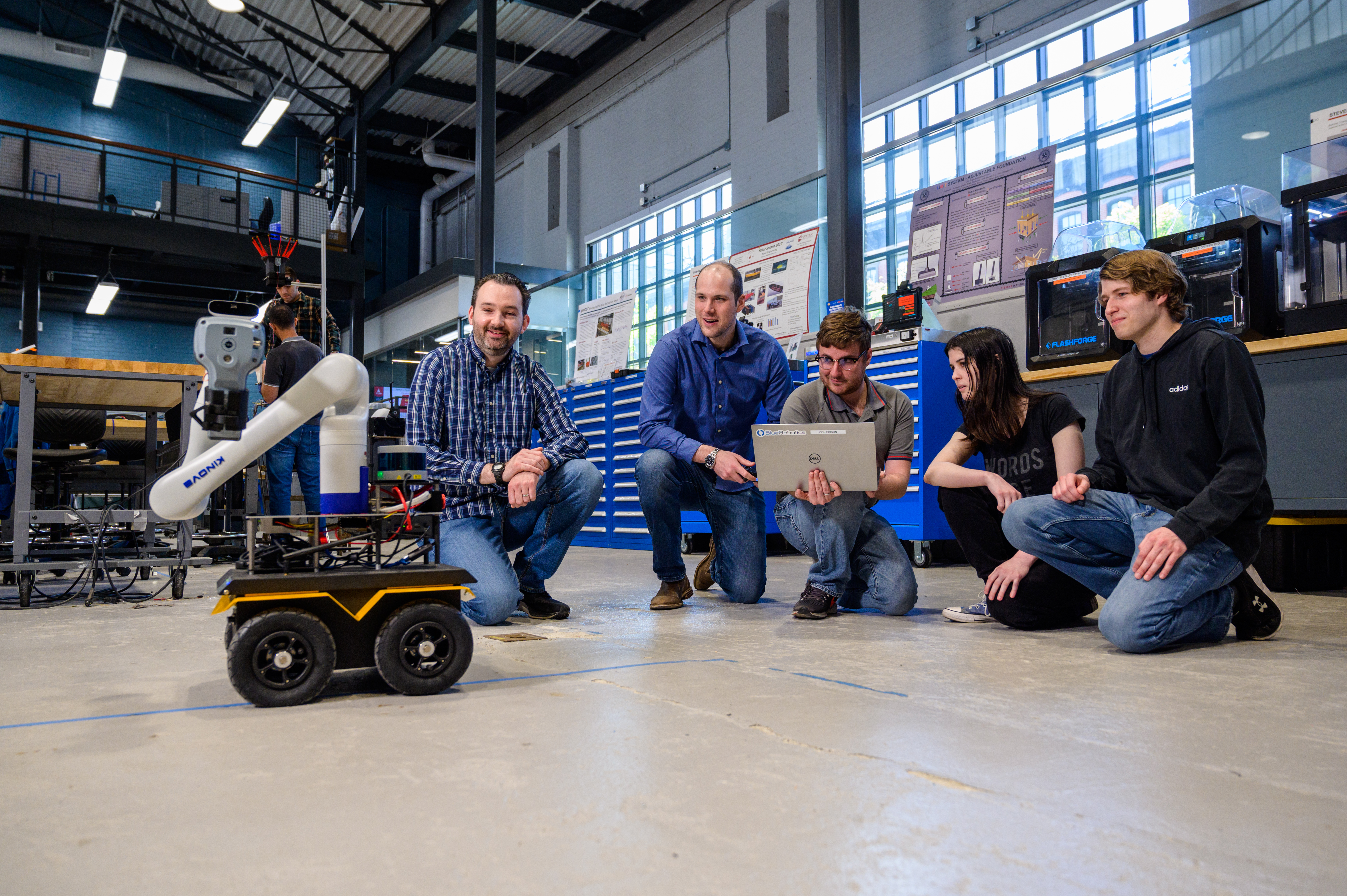 Professor Brendan Englot and a group of student kneeling down on the floor observing a laptop-controlled robot.