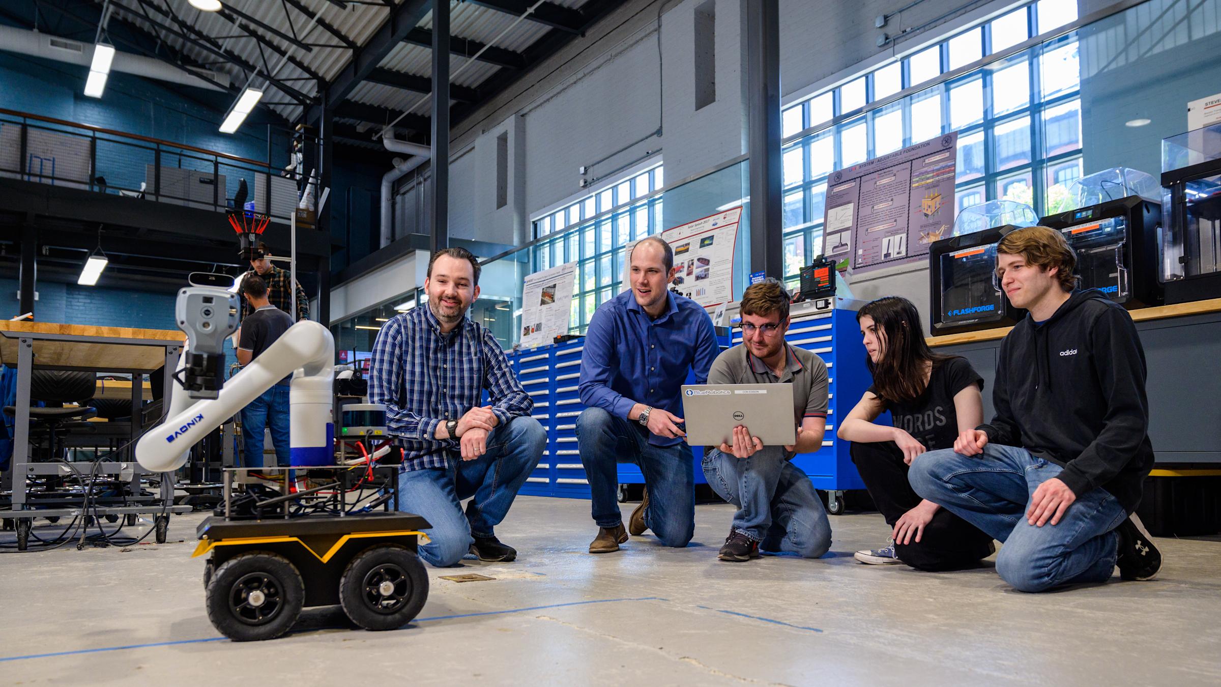 Professor Brendan Englot and a group of student kneeling down on the floor observing a laptop-controlled robot.