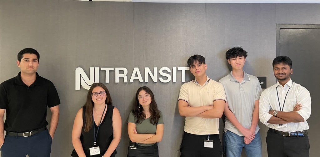 Six people stand in front of a NJ TRANSIT sign.