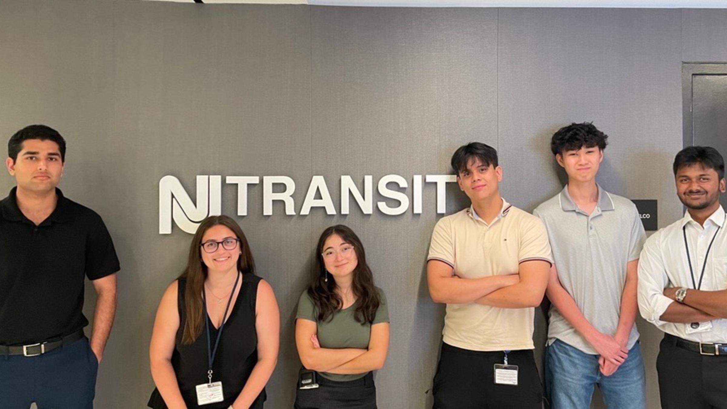 Six people stand in front of a NJ TRANSIT sign.