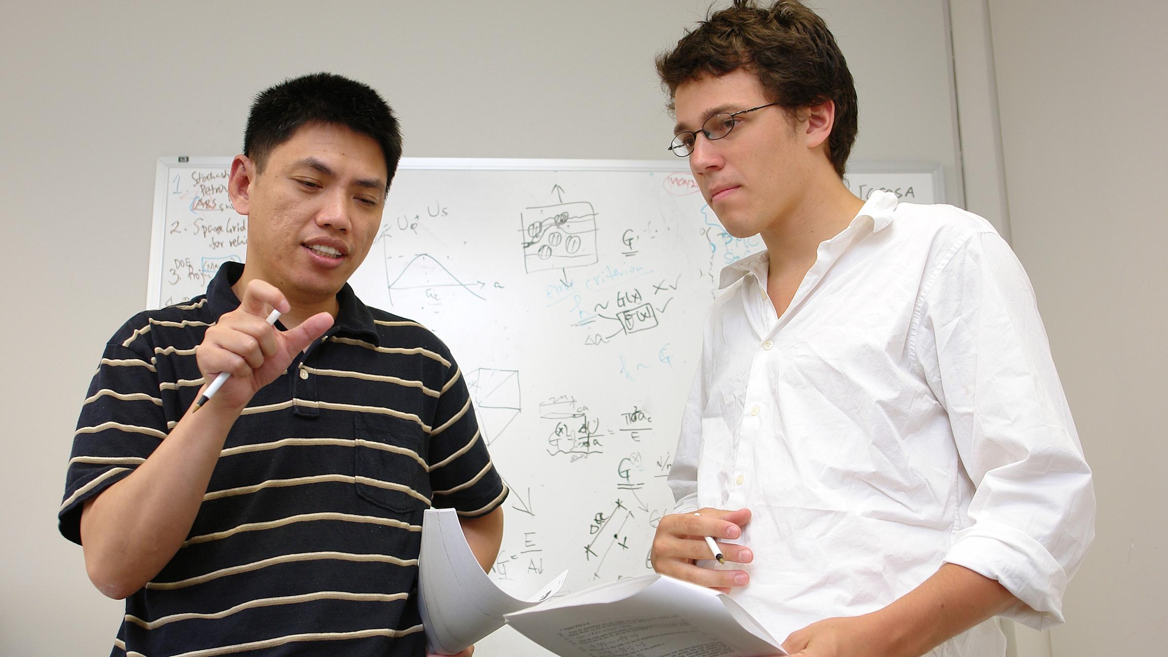 A male faculty talking to a male student in a classroom with a whiteboard in the background