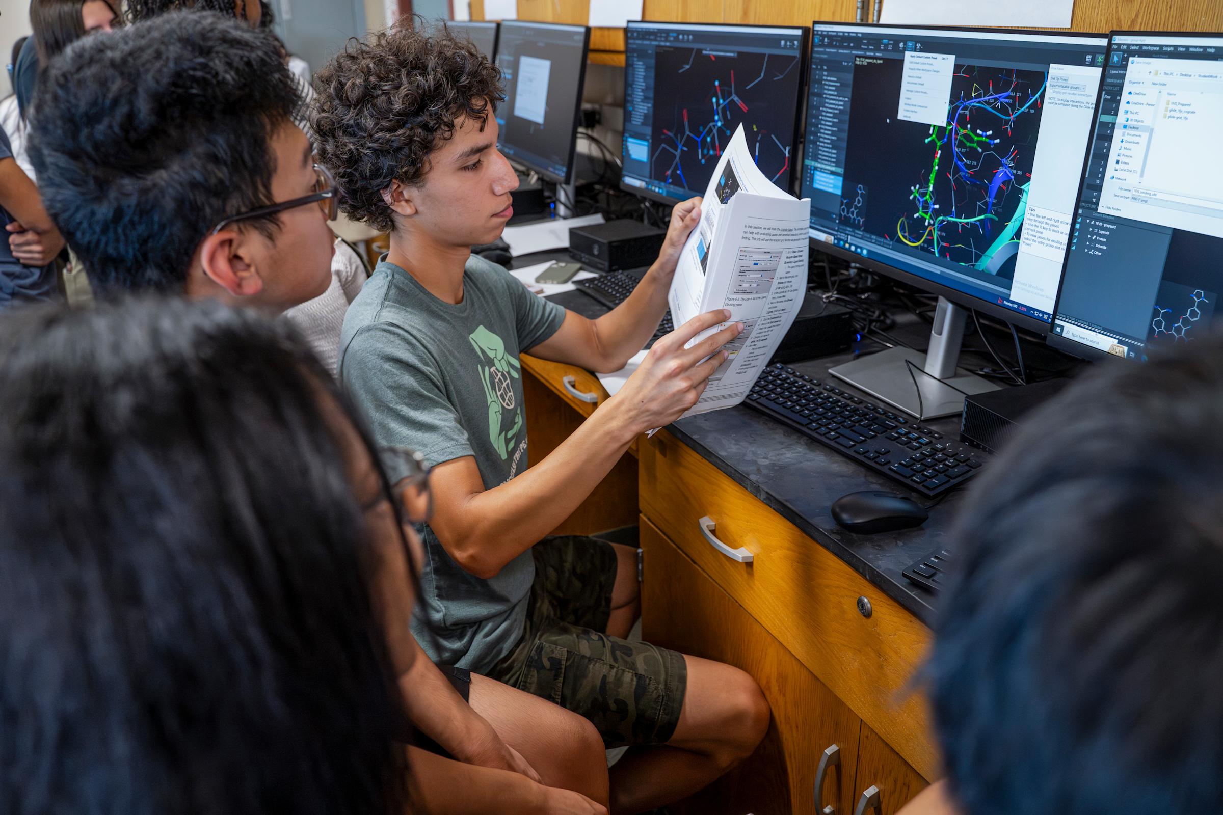 Two high school students sit in front of a computer monitor while reading a paper printout.