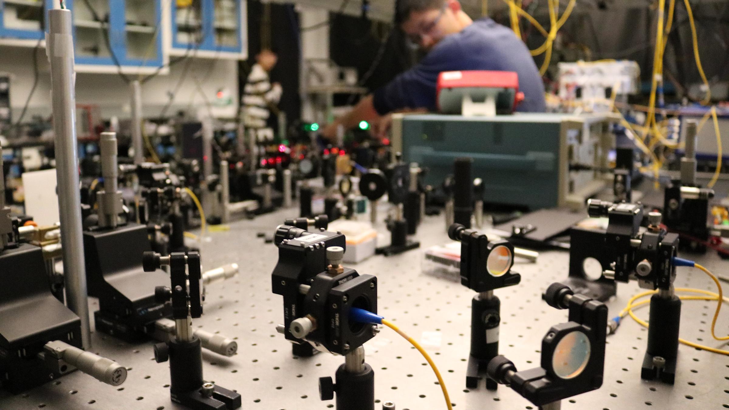 A photo of a male student working in the quantum lab surrounded by wires and photonics equipment.