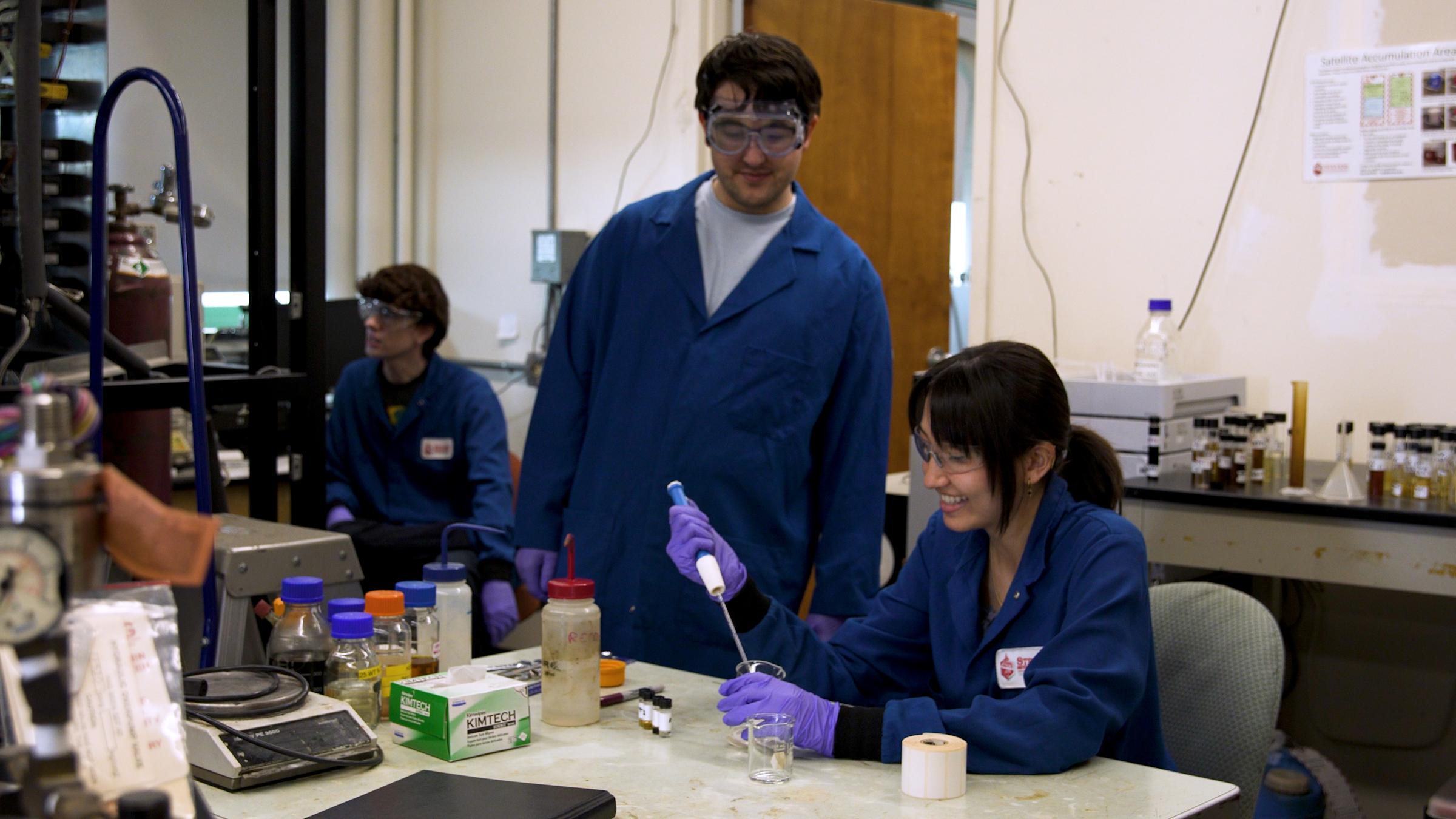 Three students in a lab conducting an experiment. From the left, a male student is sitting, a male student is standing and a female student is sitting in a chair using a syringe to conduct the experiment.