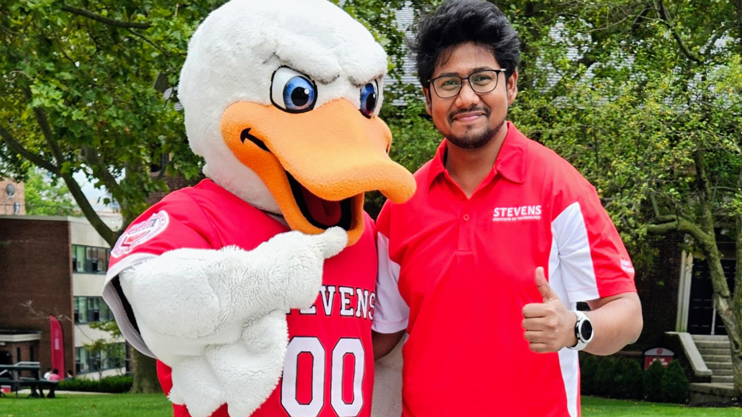A man in a Stevens polo shirt poses next to Attila, Stevens' duck mascot, while the man in the polo shirt gives a thumbs up.
