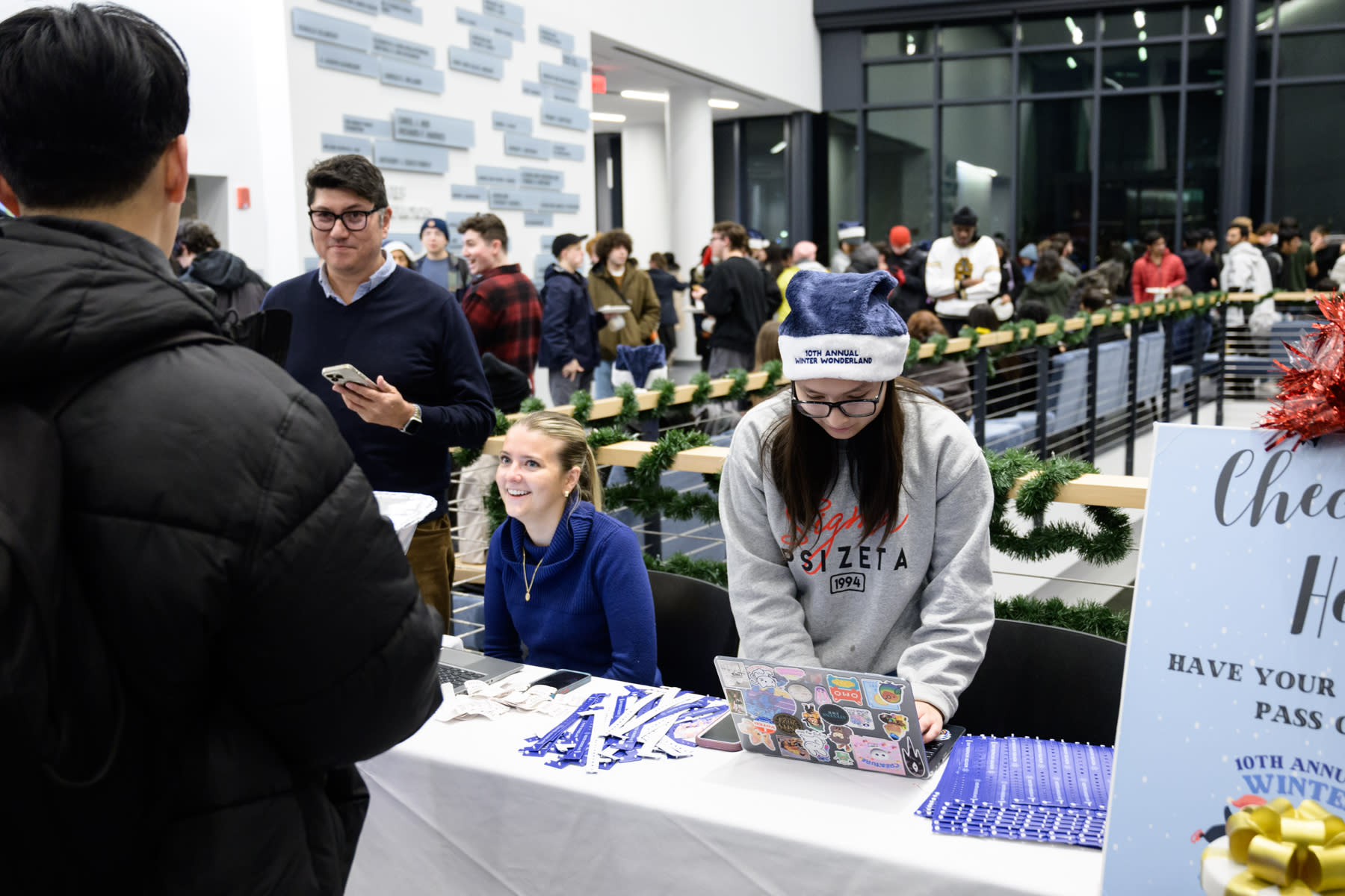 A check-in table at the Winter Wonderland event, with a few people signing in a student. The table is lined with event bracelets, ready to be handed out to attendees. In the background, students are seen mingling and enjoying the festive atmosphere.