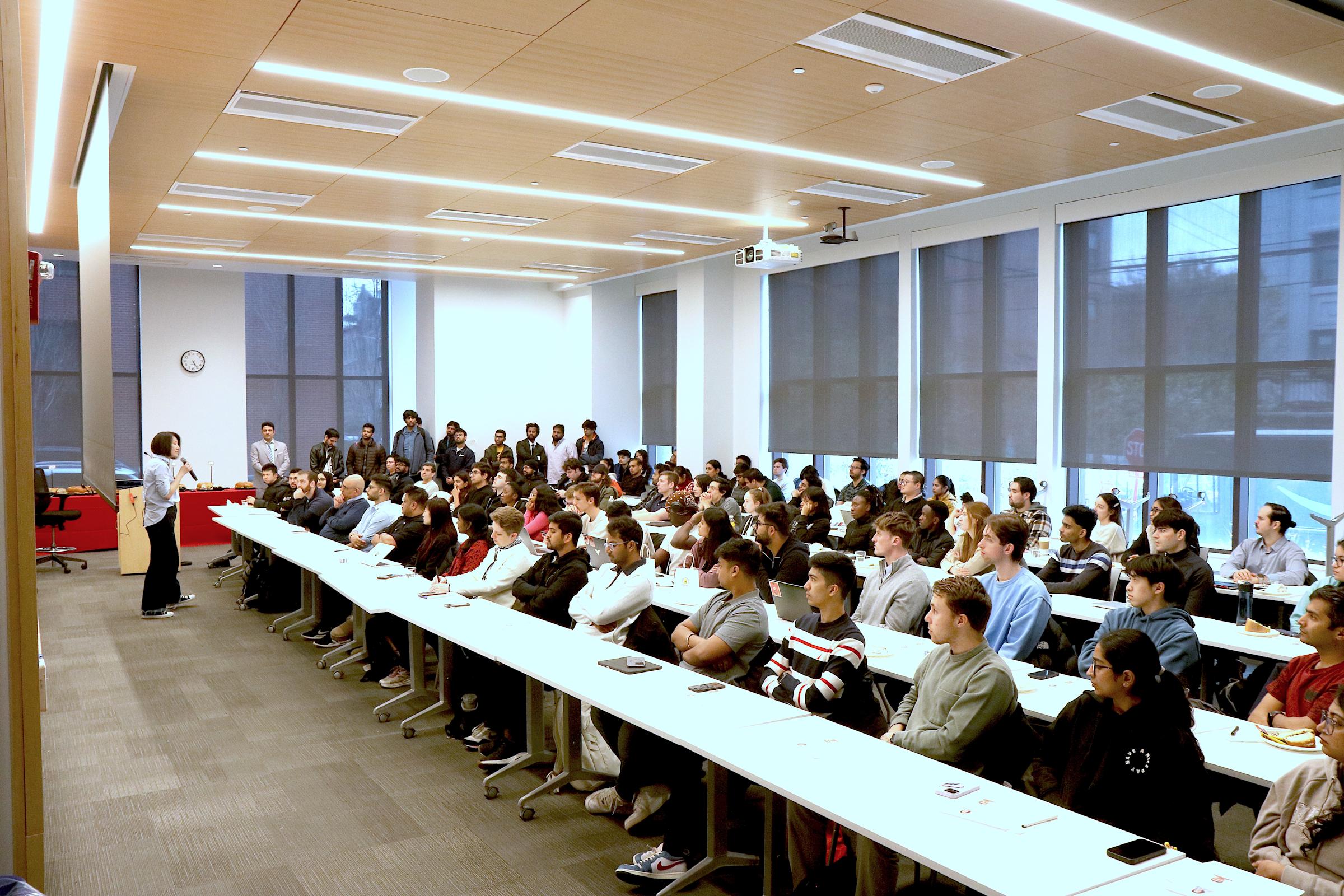 A full audience of students sit at long tables in a room looking forward at a speaker at a podium.