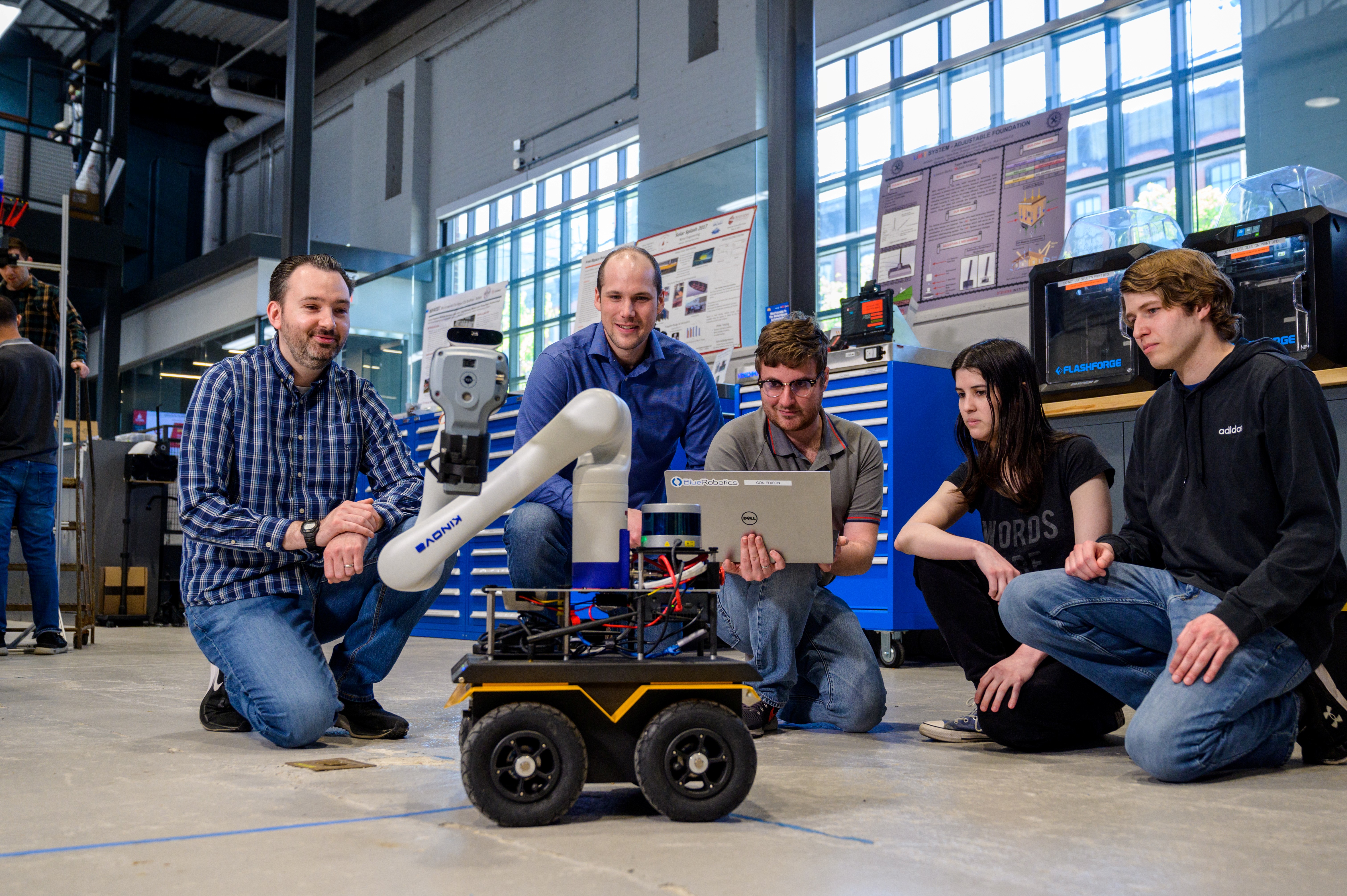 Brendan Englot, far left, kneels with four students around a wheeled mobile robot with a robotic arm in a lab. One student holds a laptop while the group observes the robot, surrounded by workshop equipment, tools and research displays.