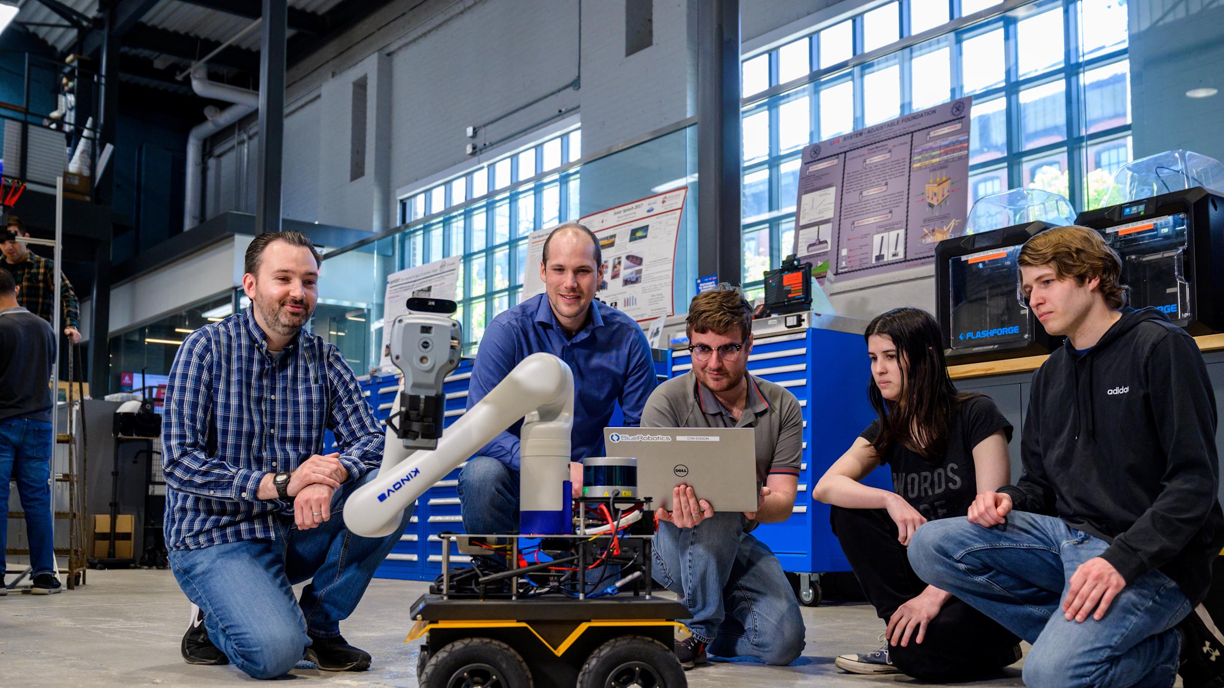 Brendan Englot, far left, kneels with four students around a wheeled mobile robot with a robotic arm in a lab. One student holds a laptop while the group observes the robot, surrounded by workshop equipment, tools and research displays.