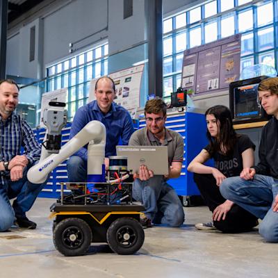 Brendan Englot, far left, kneels with four students around a wheeled mobile robot with a robotic arm in a lab. One student holds a laptop while the group observes the robot, surrounded by workshop equipment, tools and research displays.