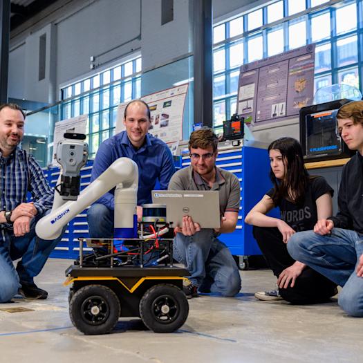 Brendan Englot, far left, kneels with four students around a wheeled mobile robot with a robotic arm in a lab. One student holds a laptop while the group observes the robot, surrounded by workshop equipment, tools and research displays.