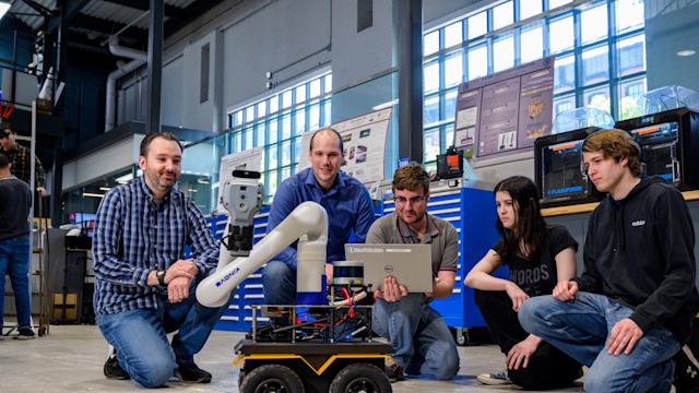Brendan Englot, far left, kneels with four students around a wheeled mobile robot with a robotic arm in a lab. One student holds a laptop while the group observes the robot, surrounded by workshop equipment, tools and research displays.