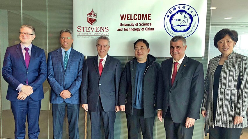 Six people in business attire stand before a banner celebrating the collaboration between Stevens and USTC.