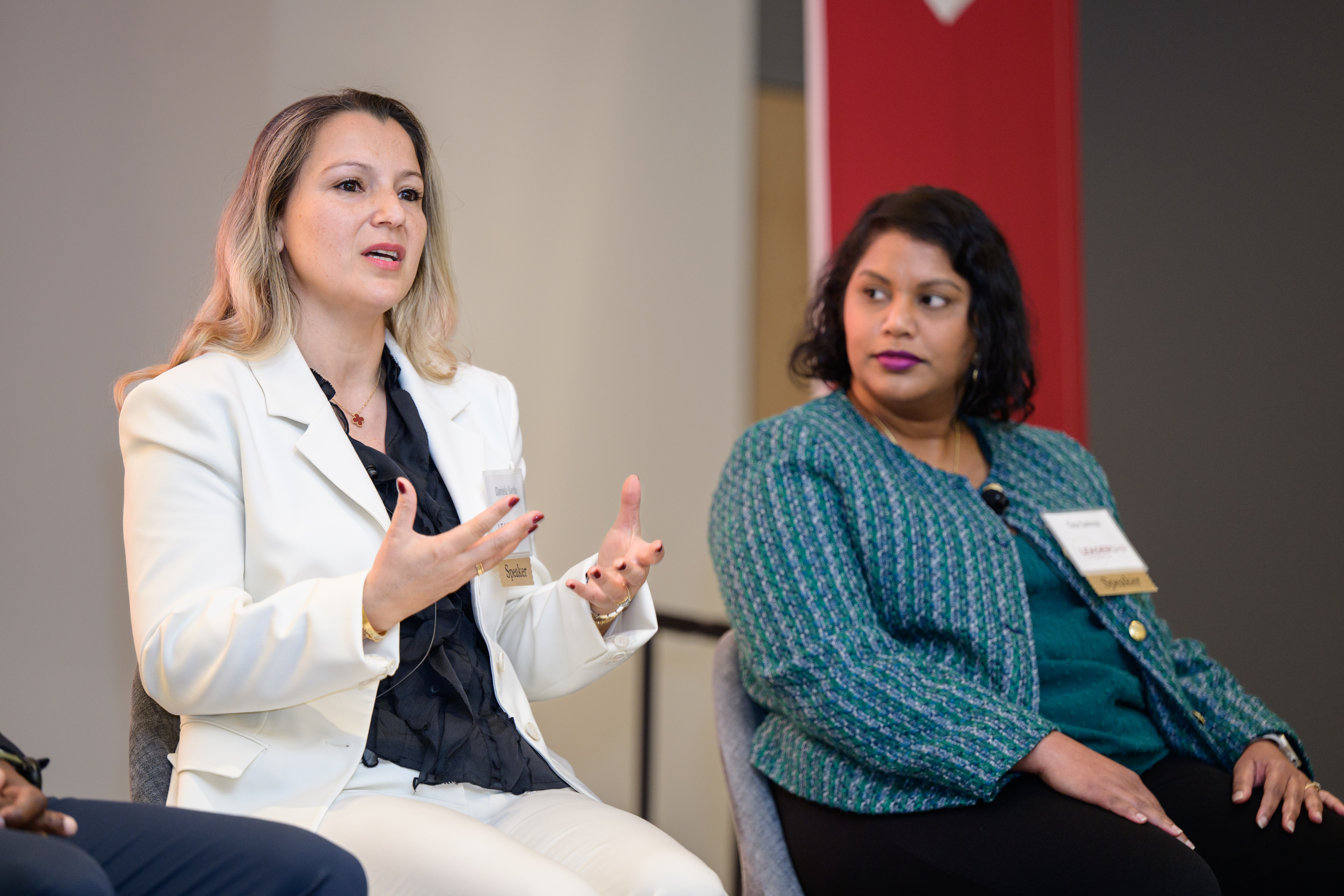 Two panelists participate in a professional networking and industry discussion. One speaker in a light-colored suit gestures while speaking, while another panelist in a green jacket listens. They are seated onstage during a professional panel event.