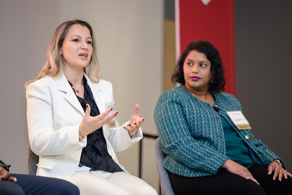 Two panelists participate in a professional networking and industry discussion. One speaker in a light-colored suit gestures while speaking, while another panelist in a green jacket listens. They are seated onstage during a professional panel event.