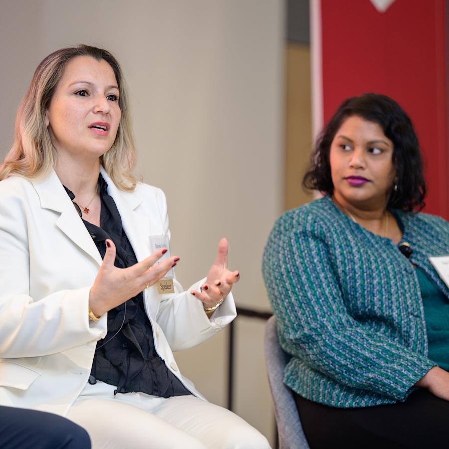 Two panelists participate in a professional networking and industry discussion. One speaker in a light-colored suit gestures while speaking, while another panelist in a green jacket listens. They are seated onstage during a professional panel event.