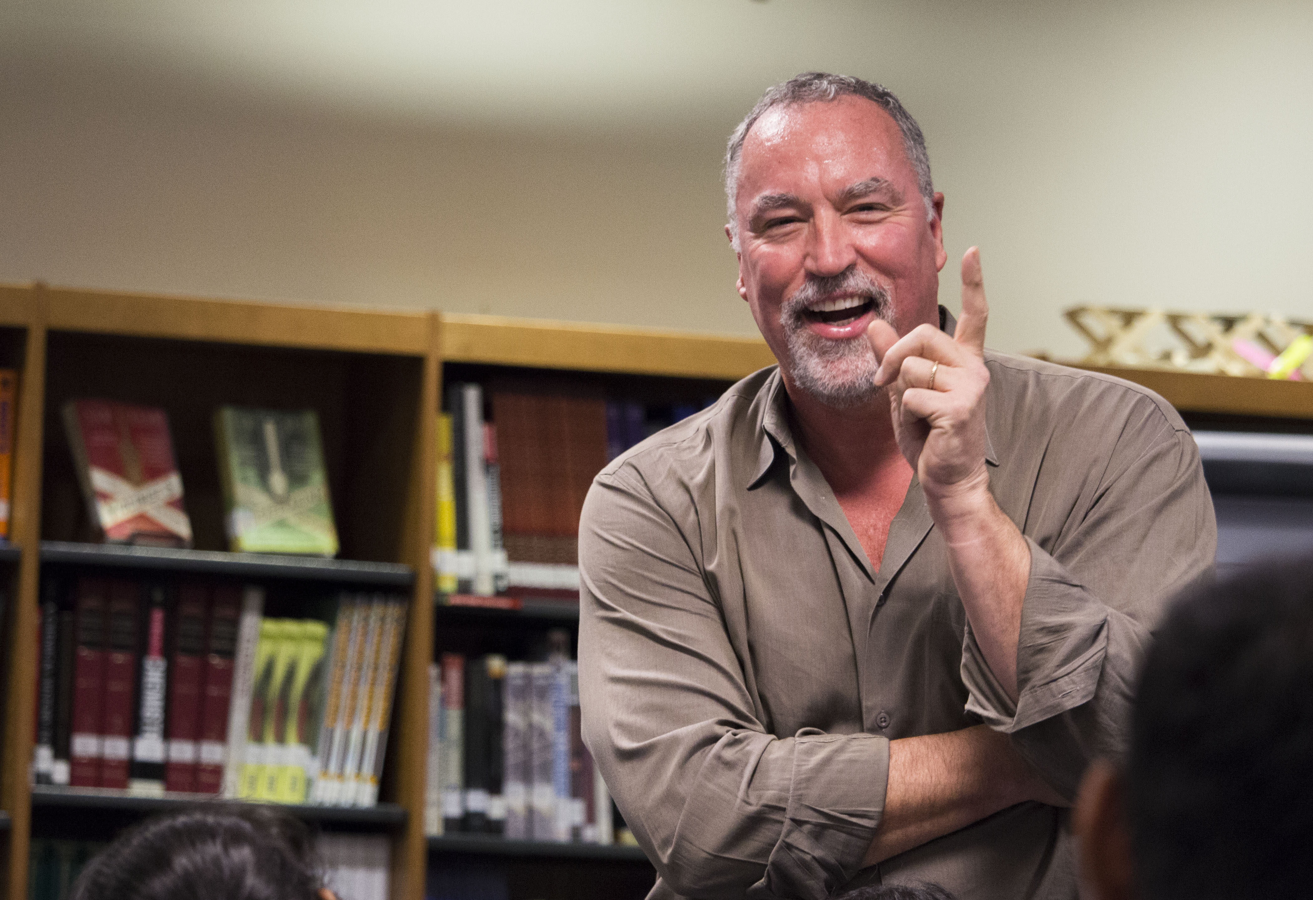 A middle-aged man with gray hair and beard wearing a tan button-down shirt smiles and gestures animatedly while sitting in an office with bookshelves in the background. The candid portrait captures him mid-conversation in a relaxed professional setting.
