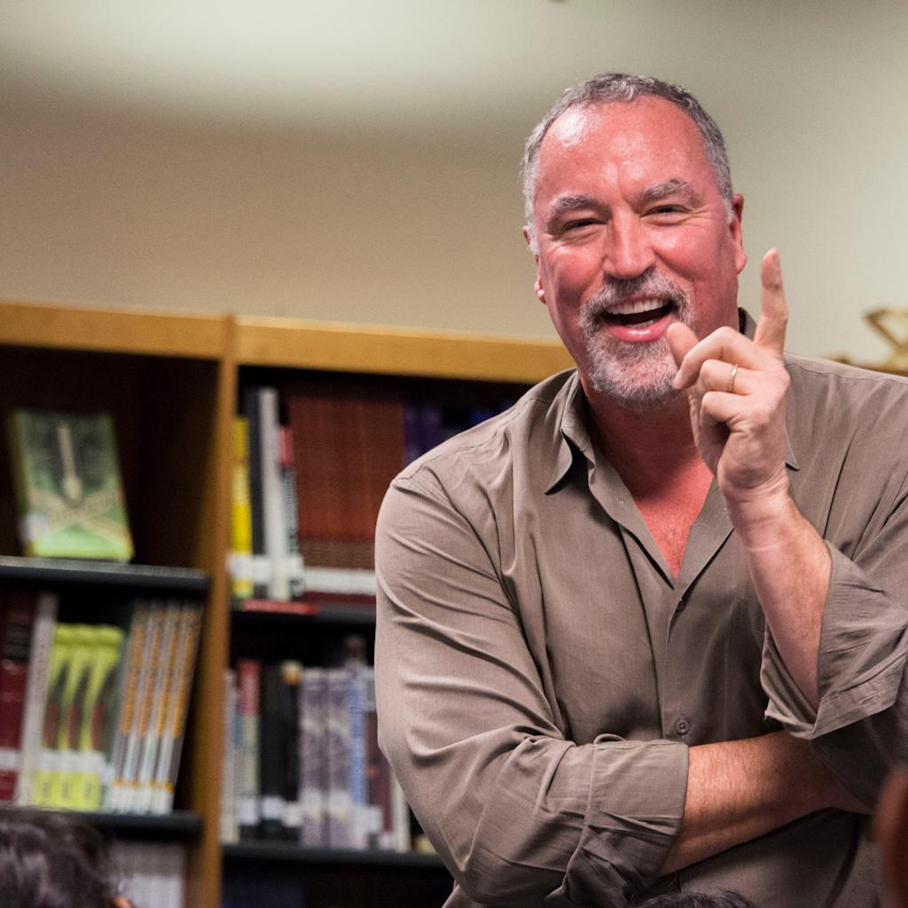 A middle-aged man with gray hair and beard wearing a tan button-down shirt smiles and gestures animatedly while sitting in an office with bookshelves in the background. The candid portrait captures him mid-conversation in a relaxed professional setting.